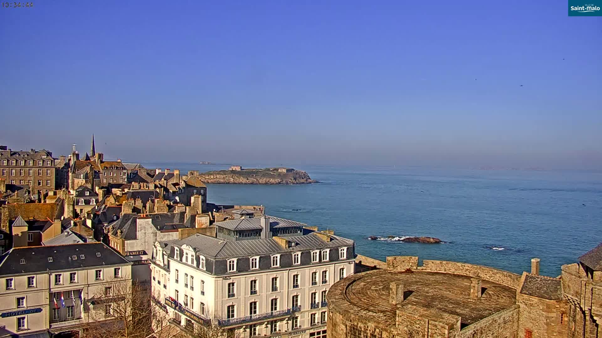 A coastal city with stone buildings and a castle overlooks a calm blue ocean and a small island under a clear blue sky.