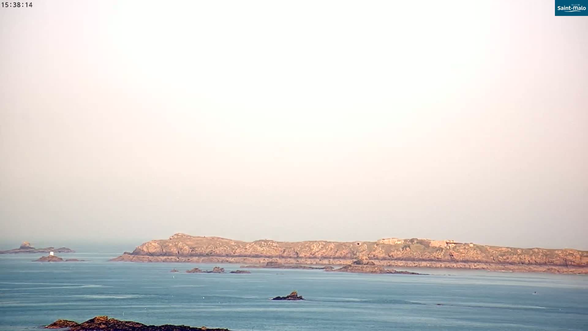 A hazy, light-colored sky overlooks a calm ocean with several rocky islands and a small islet in the distance.