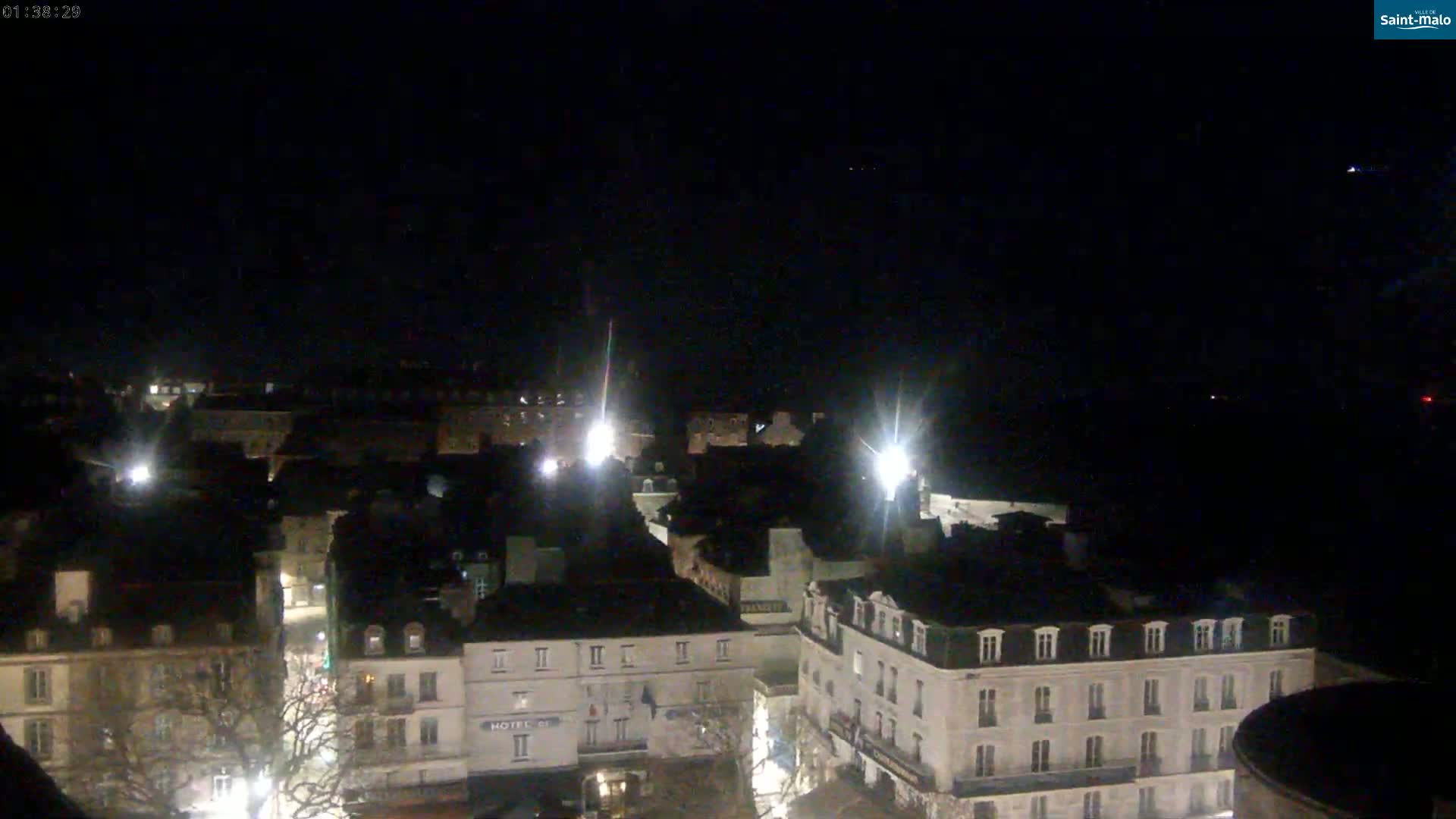 A nighttime, high-angle view shows a city square with several buildings illuminated by streetlights under a dark, clear sky.