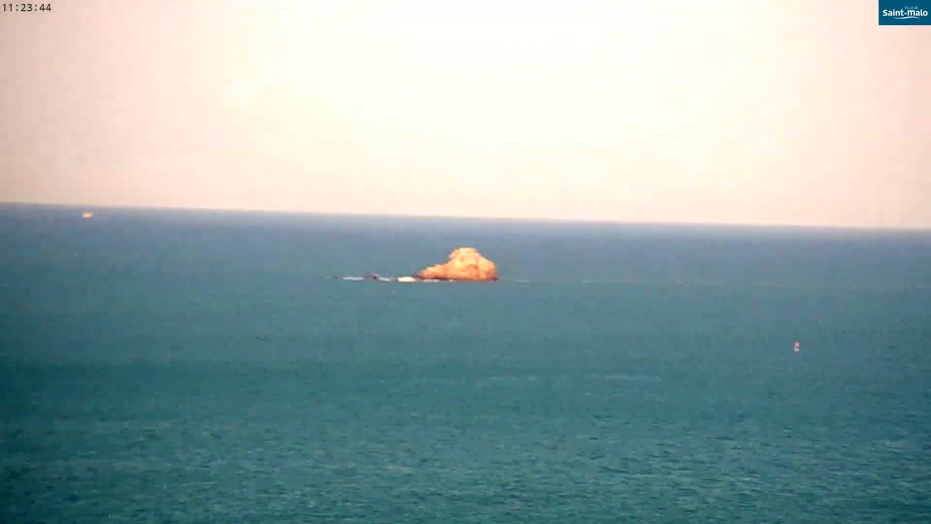 A small, light-brown rock formation sits in a calm, teal ocean under a hazy, light-pink sky.