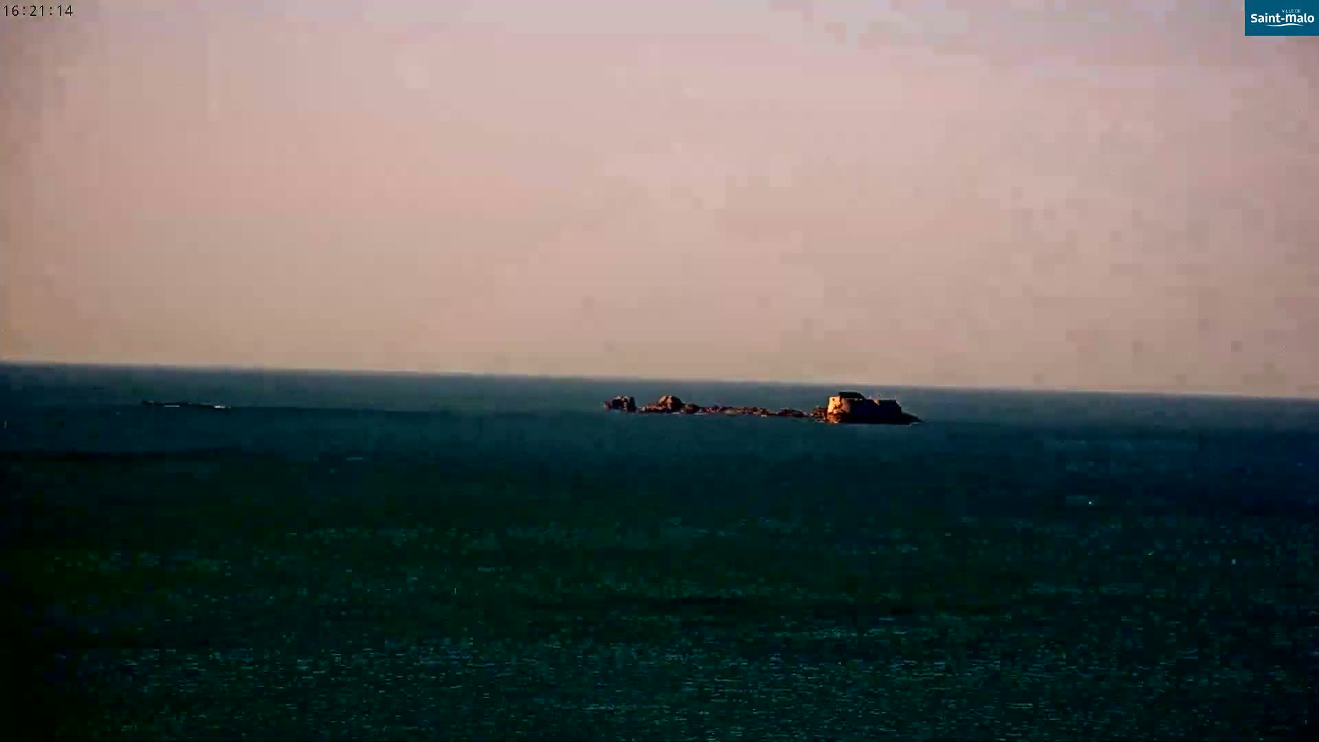 A small island with a structure on it is visible in the distance across a calm, dark-colored body of water under a pale, pinkish sky.
