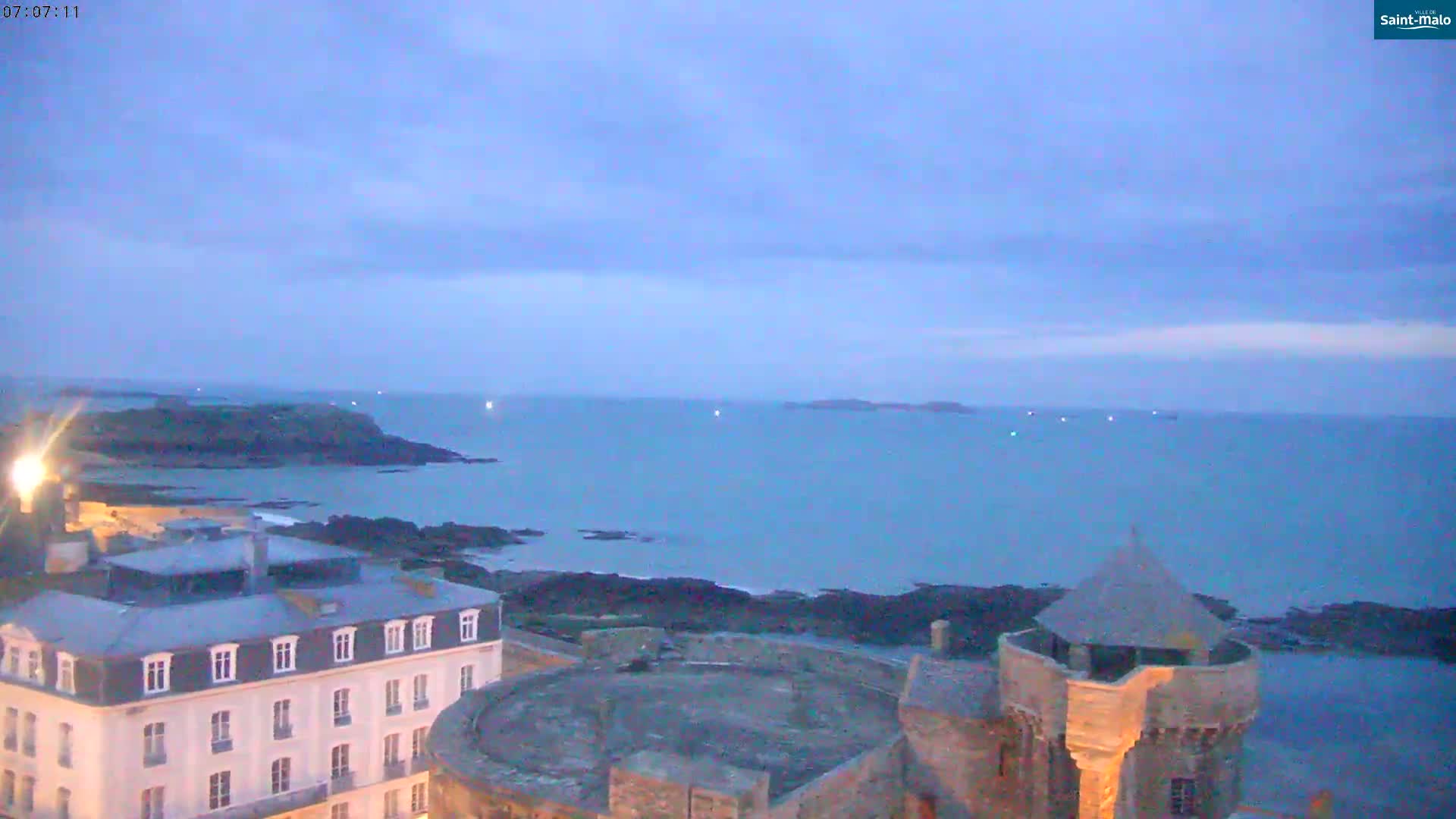 A coastal town at twilight, under a mostly clear sky, overlooks a calm sea with distant islands.