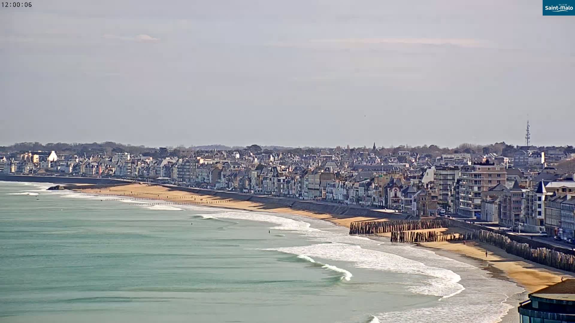 A coastal town with light-colored buildings sits next to a sandy beach with gentle waves under a mostly clear sky.