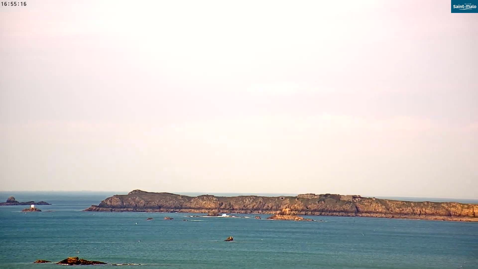 A long, low-lying rocky island is visible across a calm, blue sea under a pale, overcast sky.