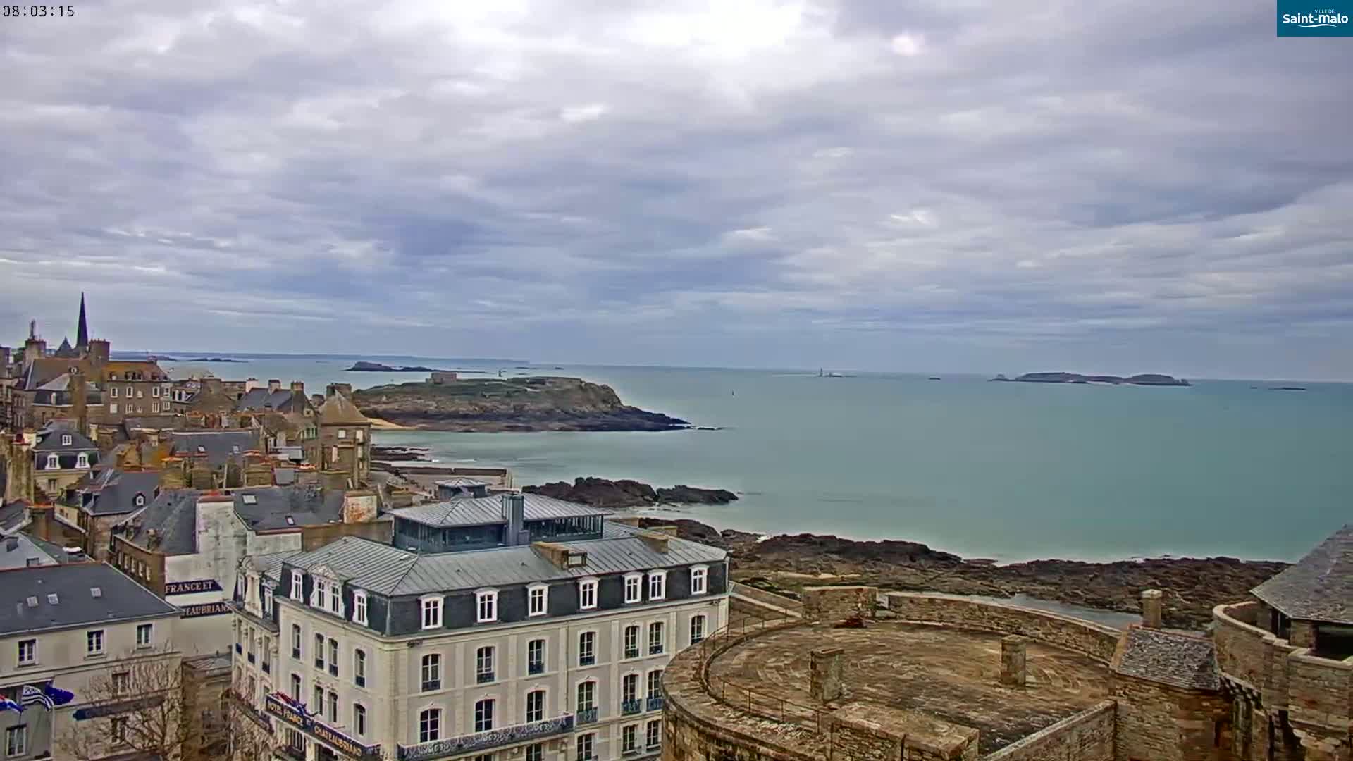 A coastal town with stone buildings overlooks a calm, teal sea under a cloudy sky.
