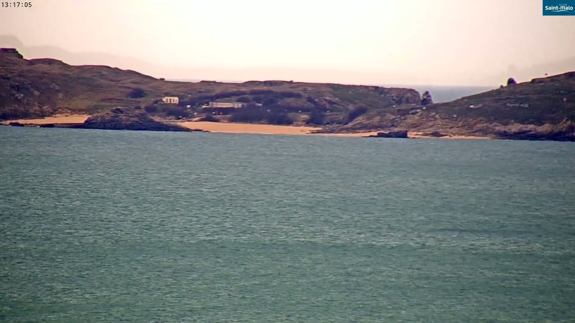 A sandy beach nestled between two rocky headlands is visible across a calm, greenish-blue body of water under a hazy sky.