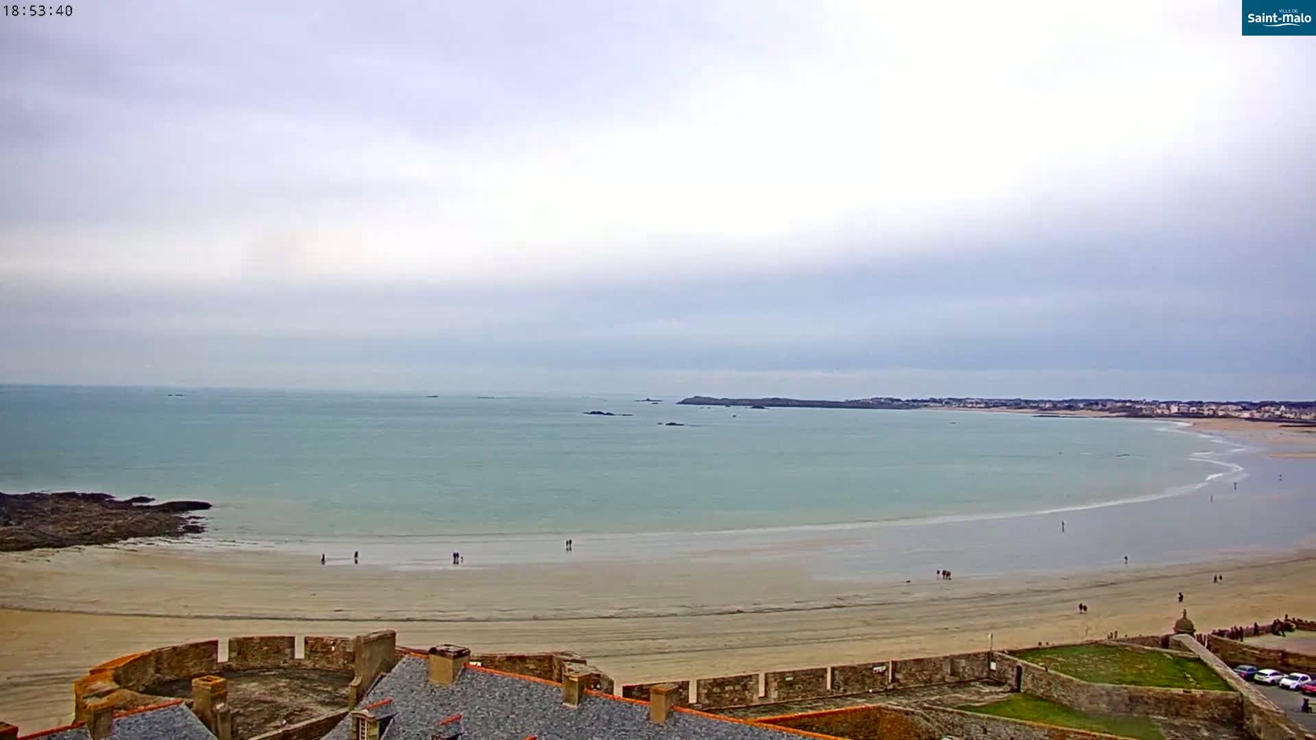 A wide sandy beach curves along a calm, light teal ocean under an overcast sky, with a stone wall and buildings in the foreground.