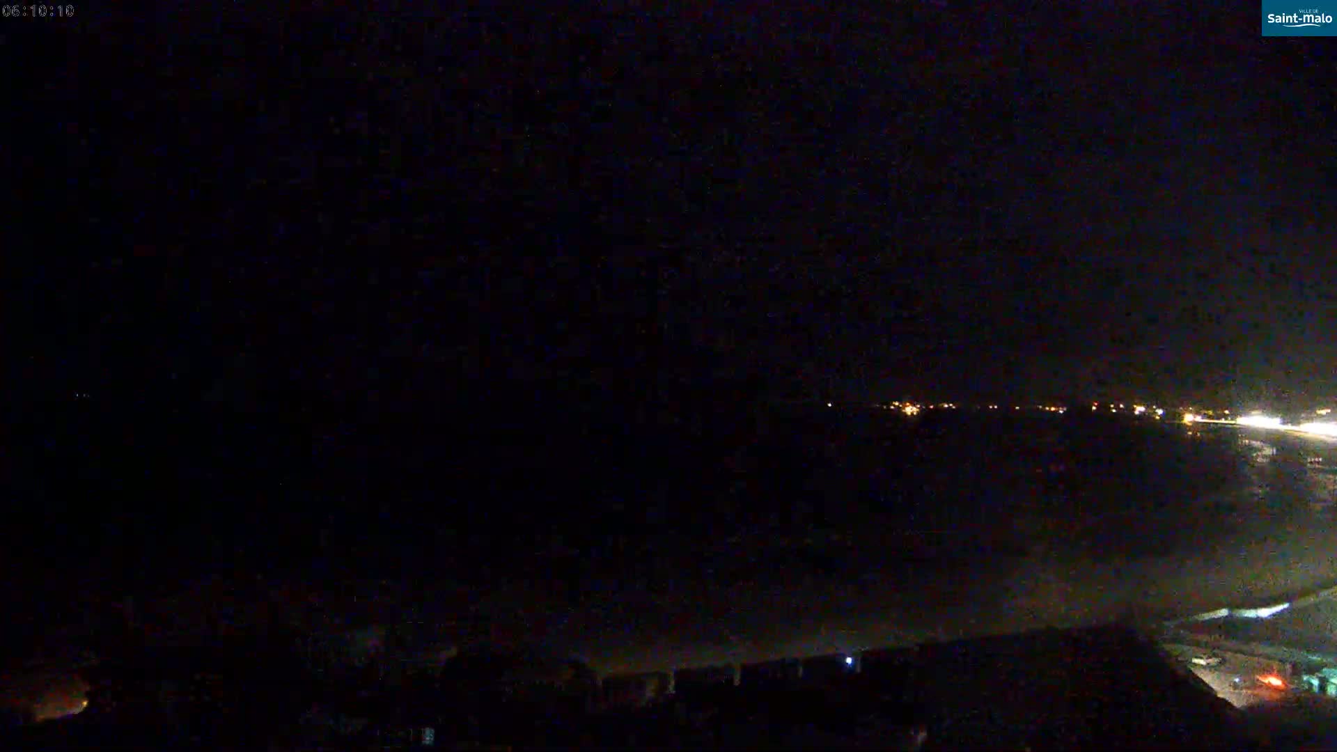 A dark nighttime scene shows a distant line of lights and a hazy foreground under a mostly clear sky.