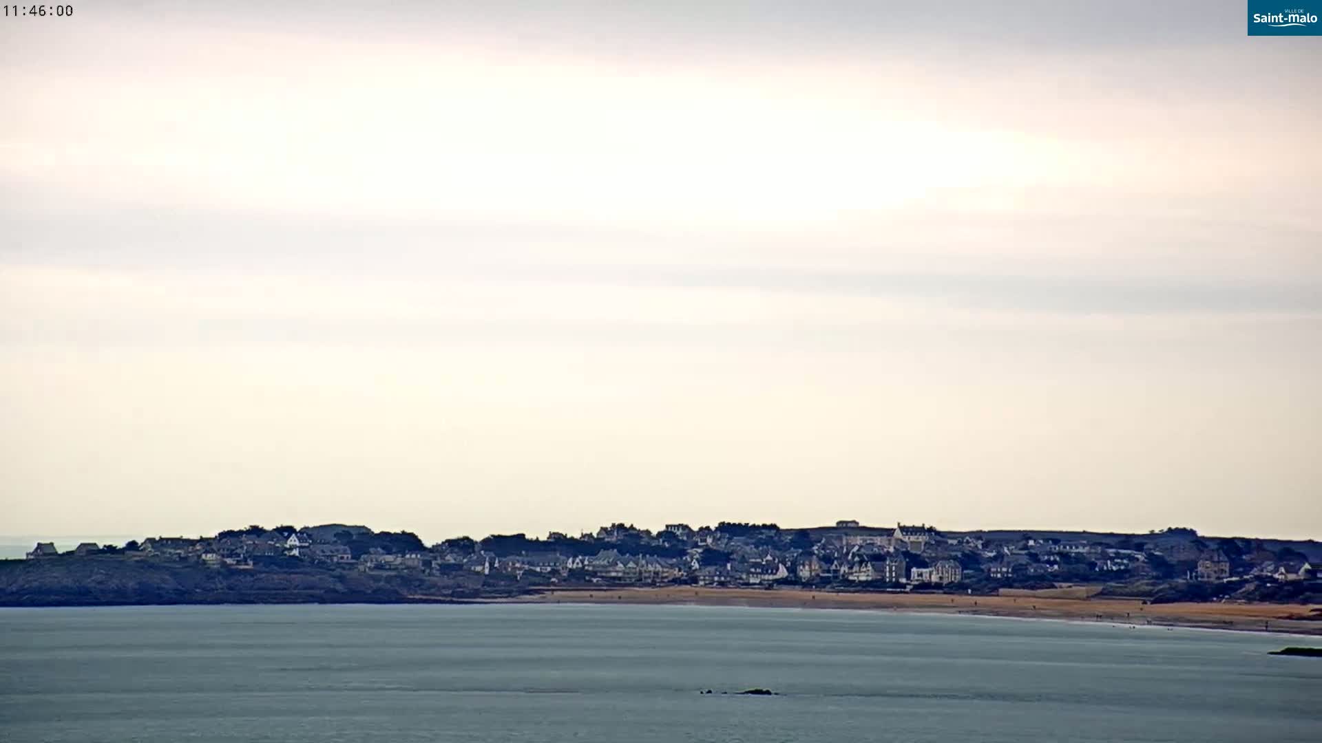 A coastal town sits on a hill overlooking a calm, gray sea under an overcast sky.