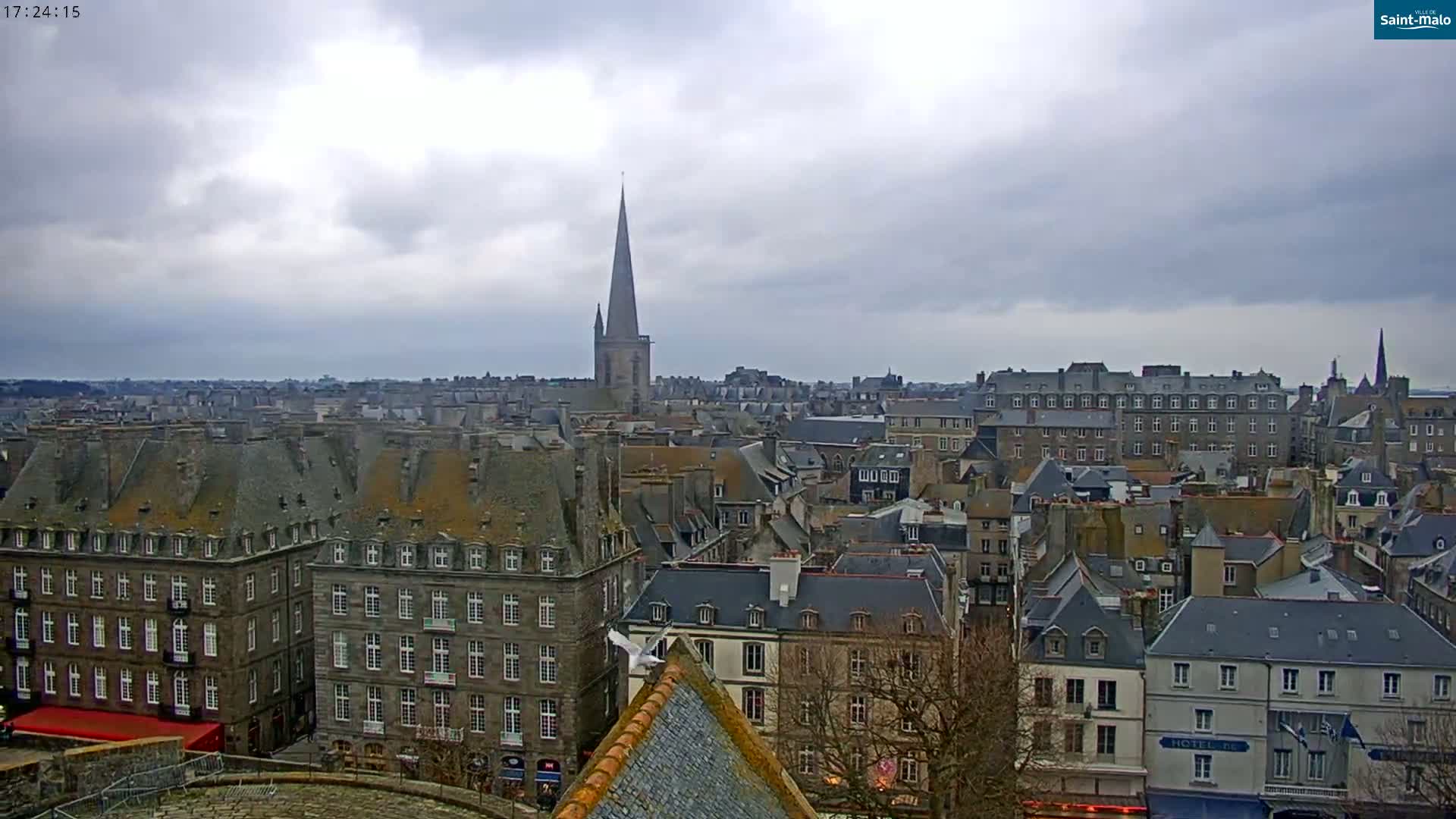 A sprawling cityscape of grey stone buildings under a cloudy sky, featuring a tall church spire.