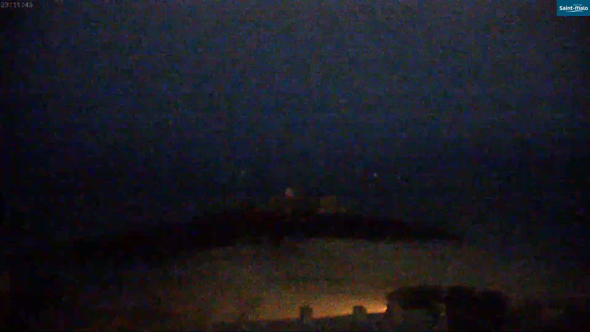 A nighttime, dark view shows a hilltop with faint lights in the distance under a mostly clear, dark sky.