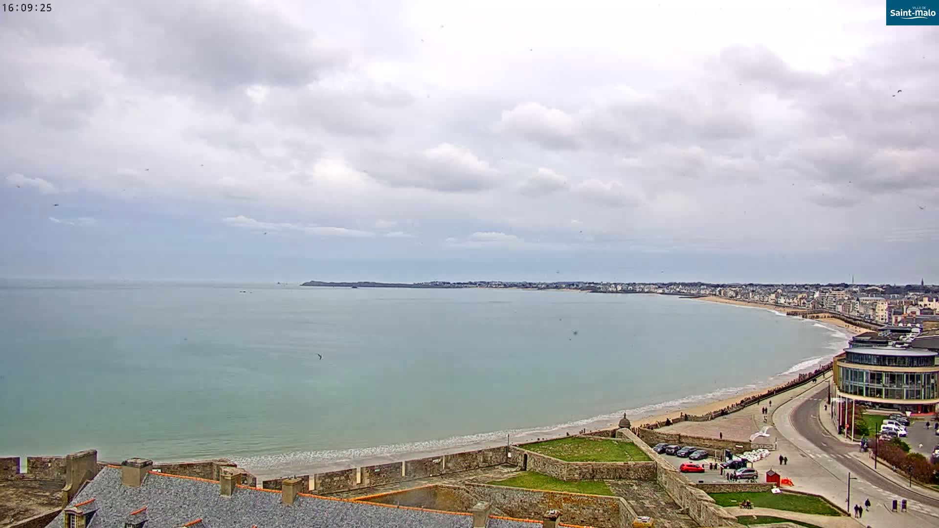 A coastal town with a sandy beach stretches along a calm, light-green ocean under a mostly cloudy sky.