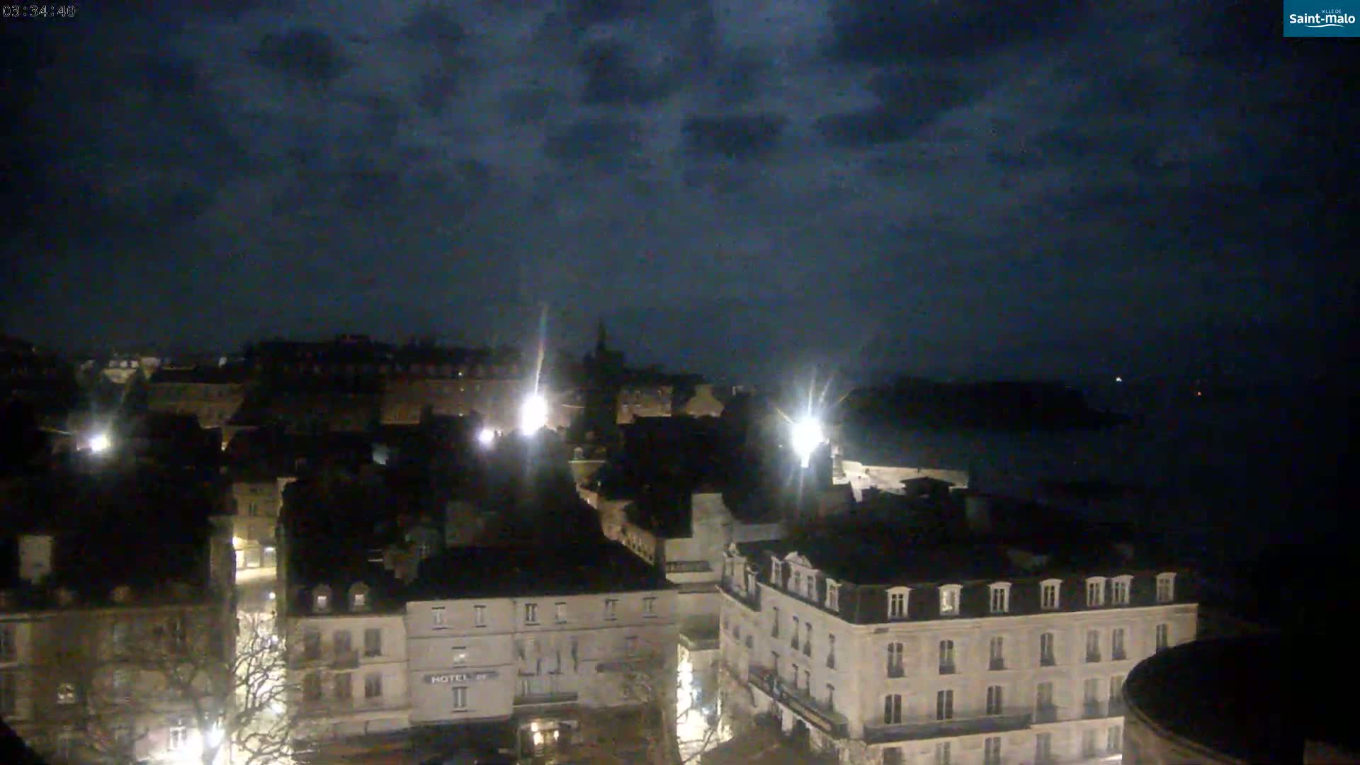 A nighttime view of a city with dark, cloudy skies, showing a collection of buildings illuminated by streetlights, and a body of dark water in the distance.