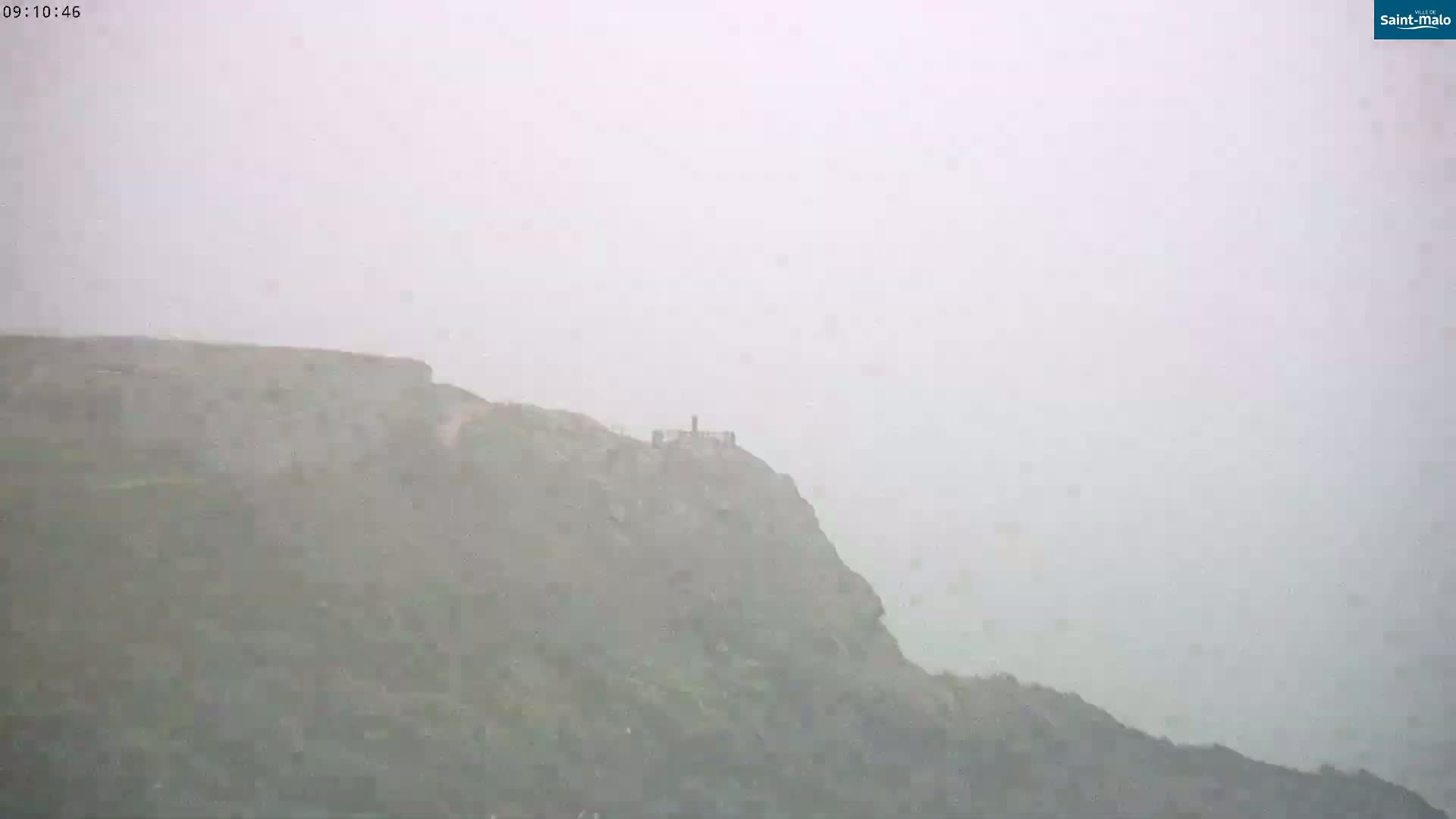 A hazy, possibly snowy or rainy, view of a cliff with a small structure atop it.