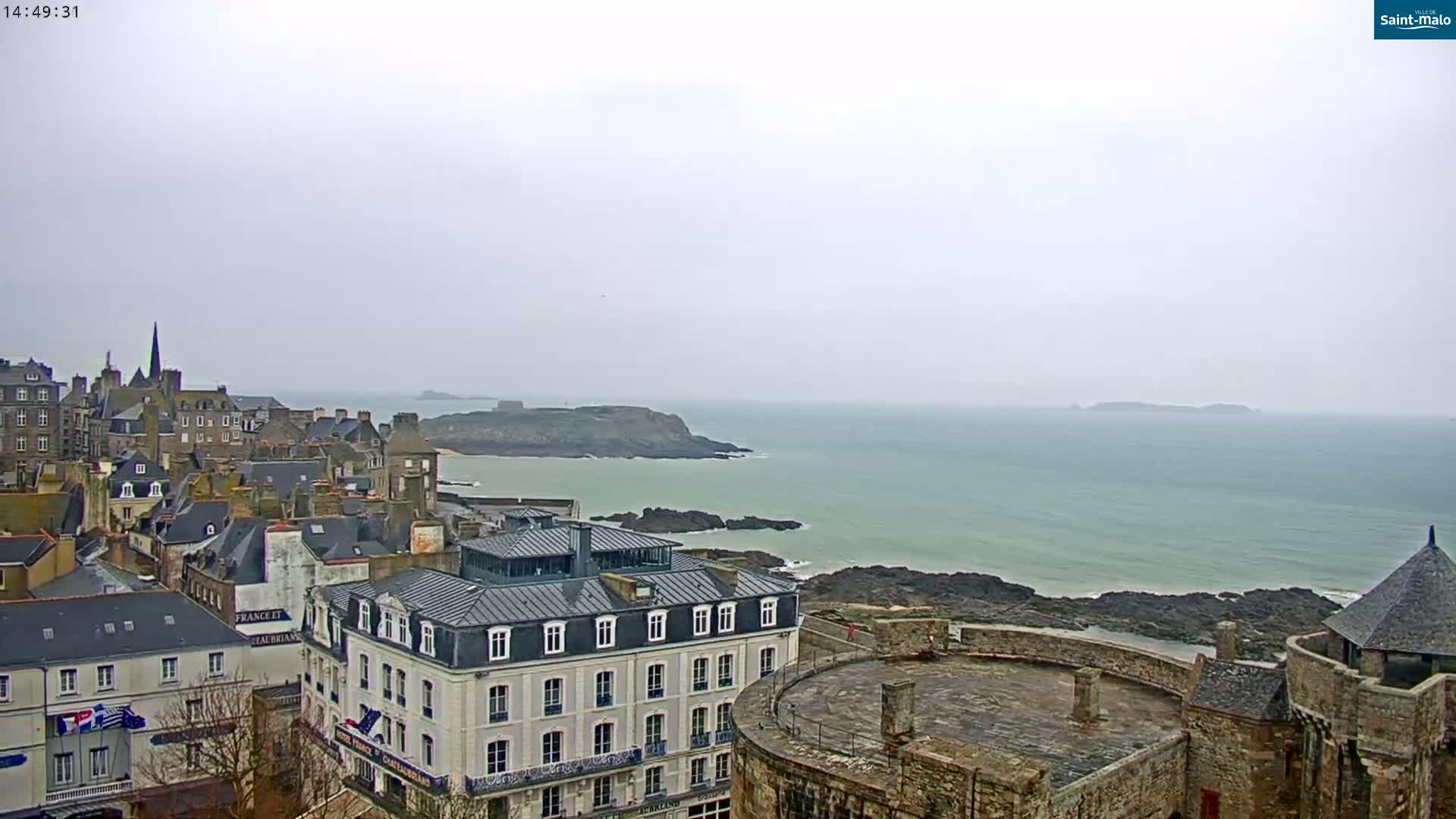 A coastal town with stone buildings and a partially visible castle overlooks a calm, gray sea under an overcast sky.