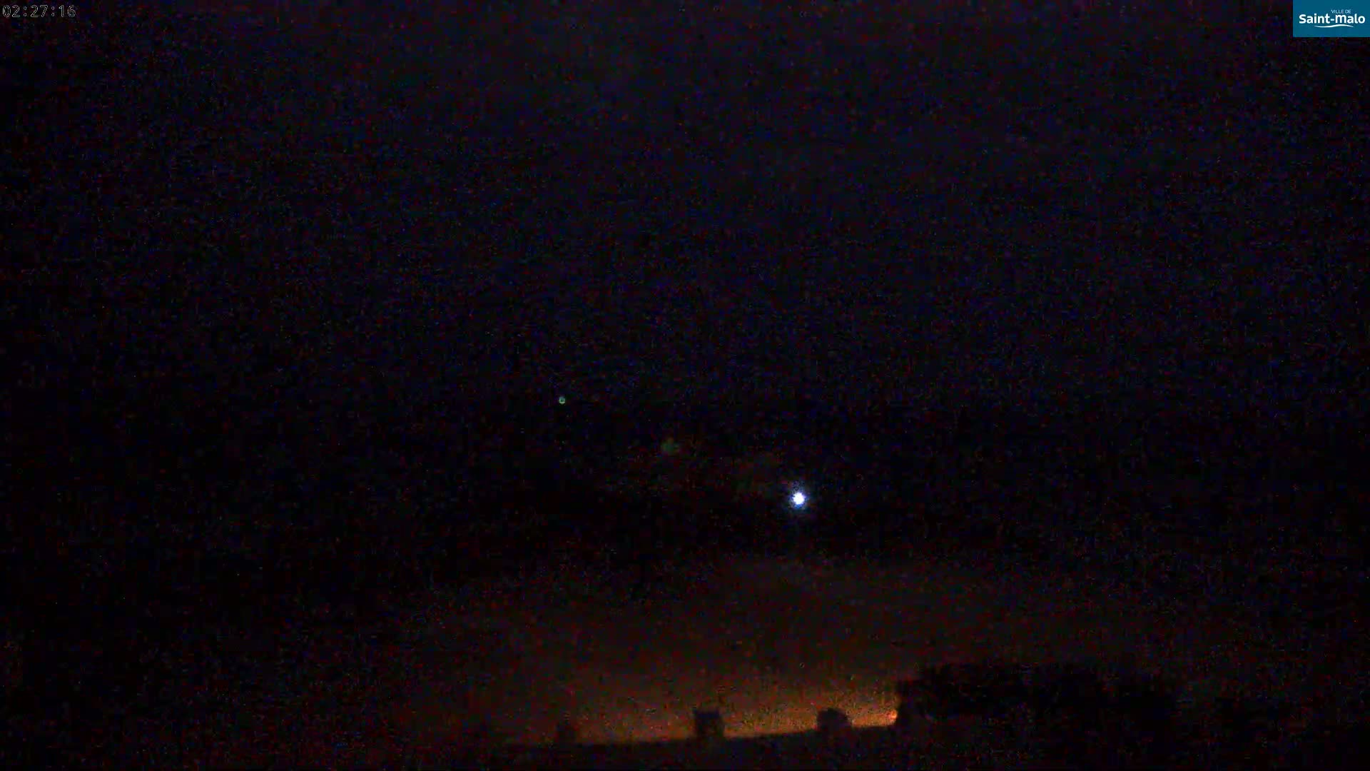A night scene shows a dark, cloudy sky with a few faint lights visible in the distance, and a brighter light possibly a streetlight, closer to the horizon.