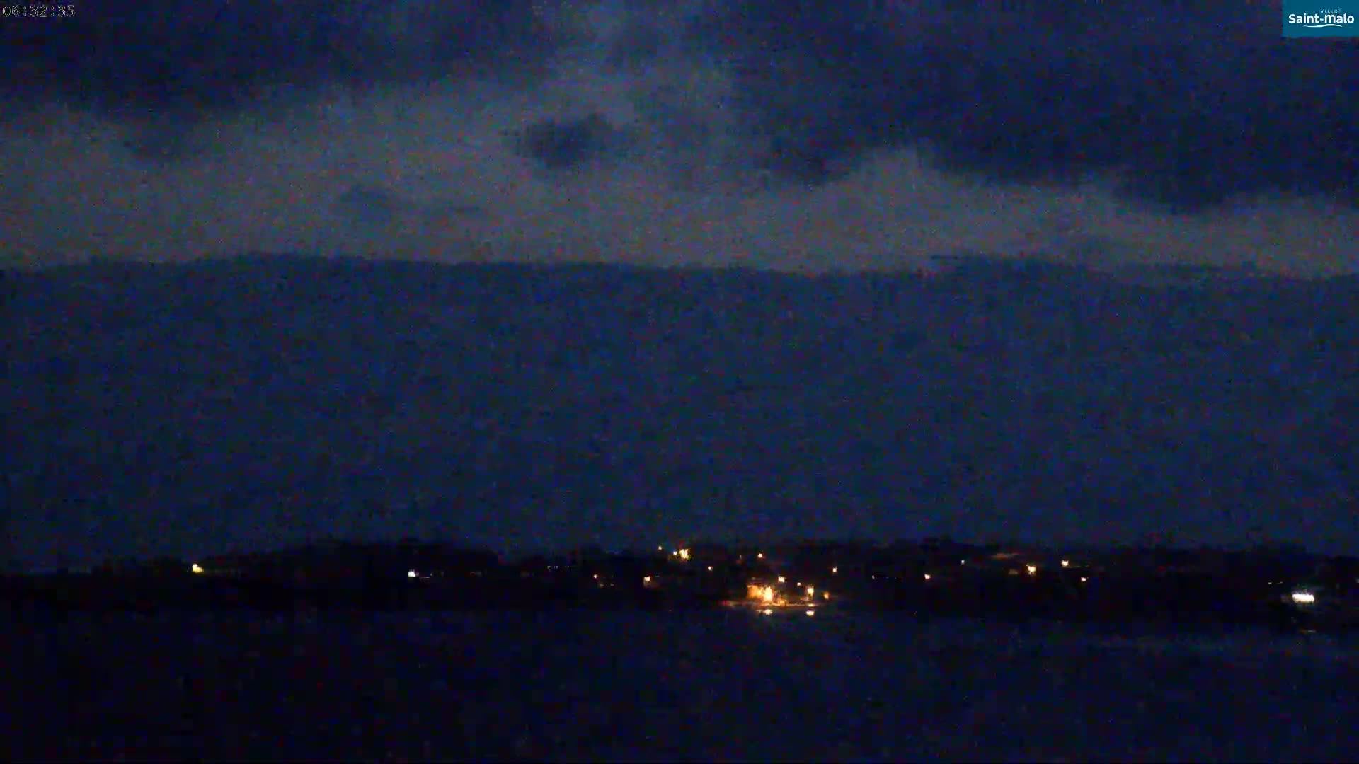 A dark, nighttime scene shows a body of water in the foreground with a line of dimly lit land in the distance under a mostly cloudy sky.
