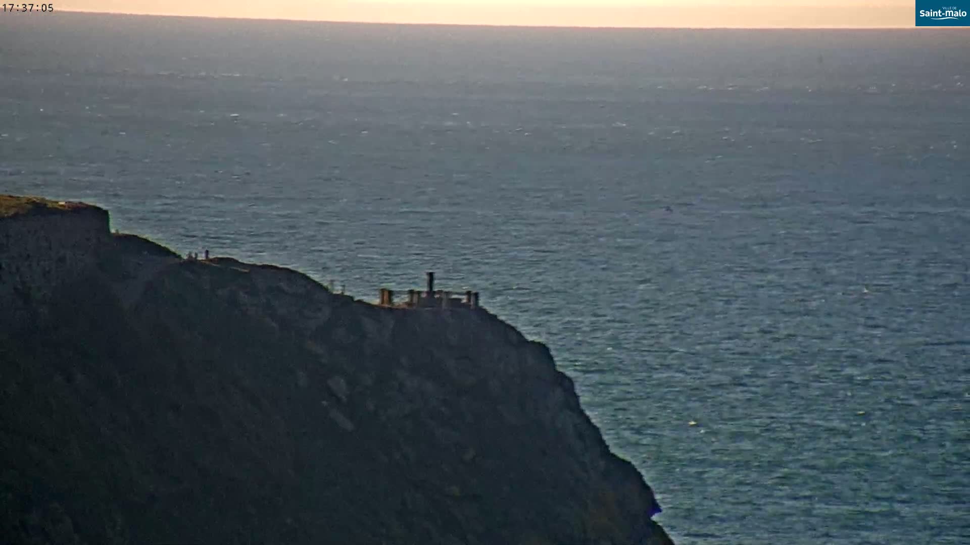 A dark cliff overlooking a calm ocean at sunset, with a small structure atop the cliff.