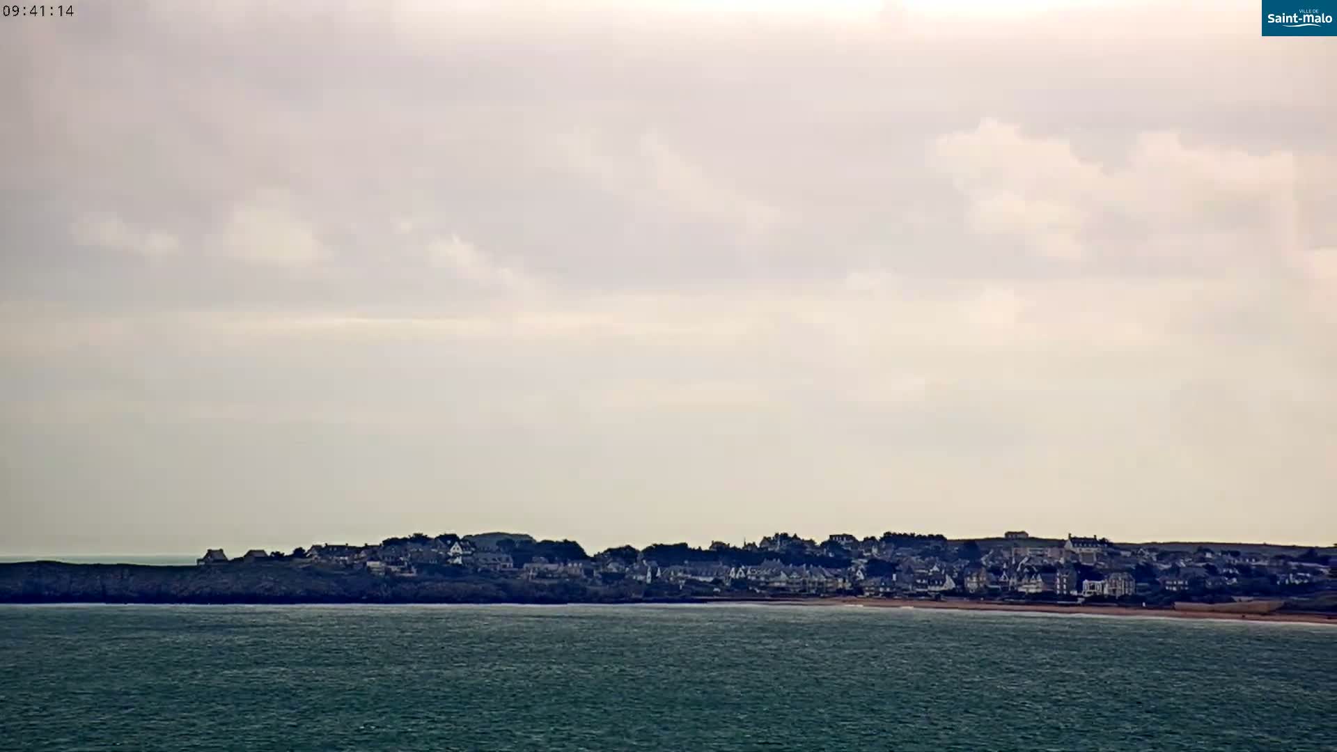 A coastal town sits on a cliff overlooking a calm, dark-green sea under a mostly cloudy sky.
