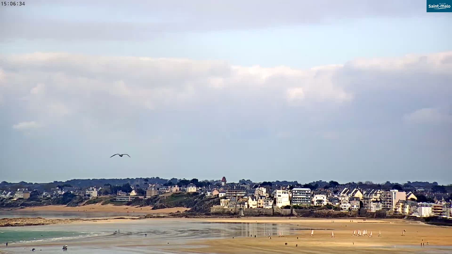 A coastal town is visible across a wide sandy beach with calm water under a partly cloudy sky, and a single bird is flying overhead.