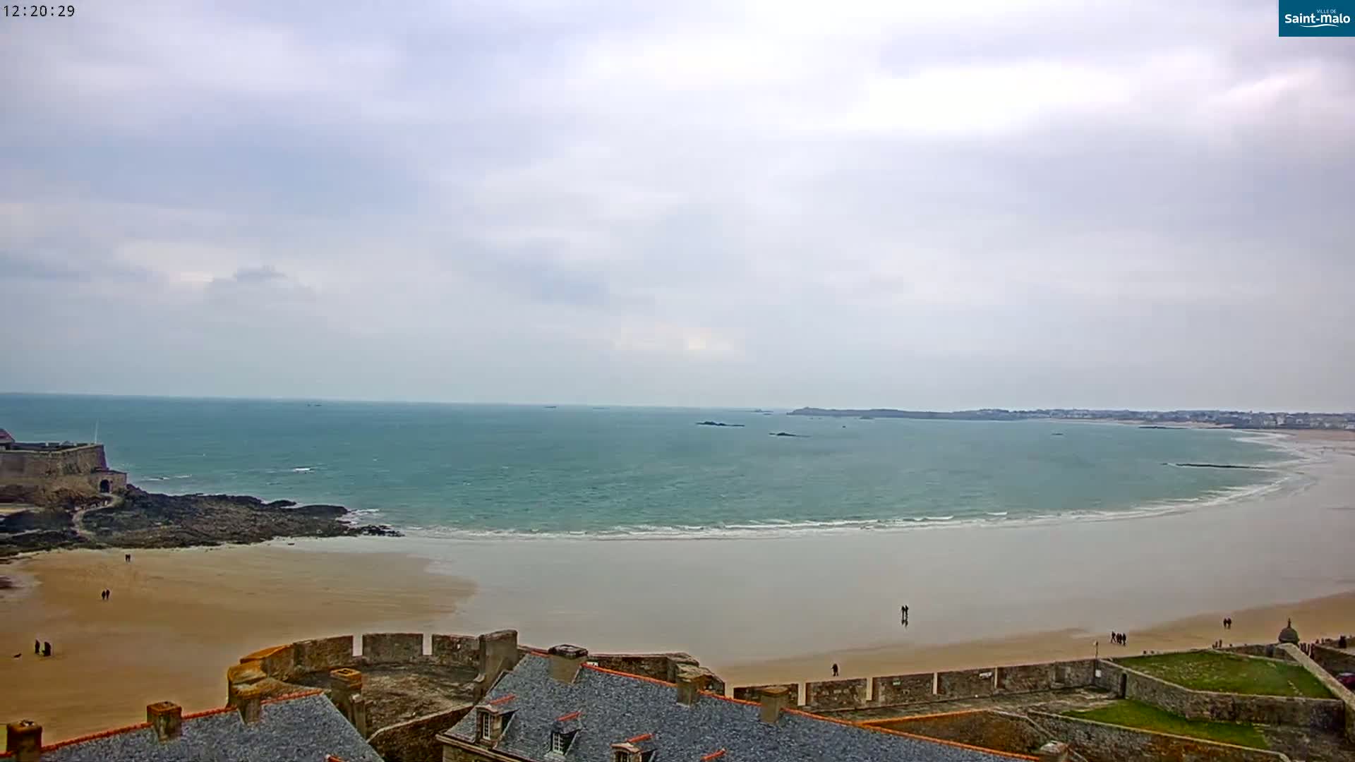 A coastal scene shows a sandy beach curving around a body of water under an overcast sky, with a stone fortress and buildings visible in the foreground.