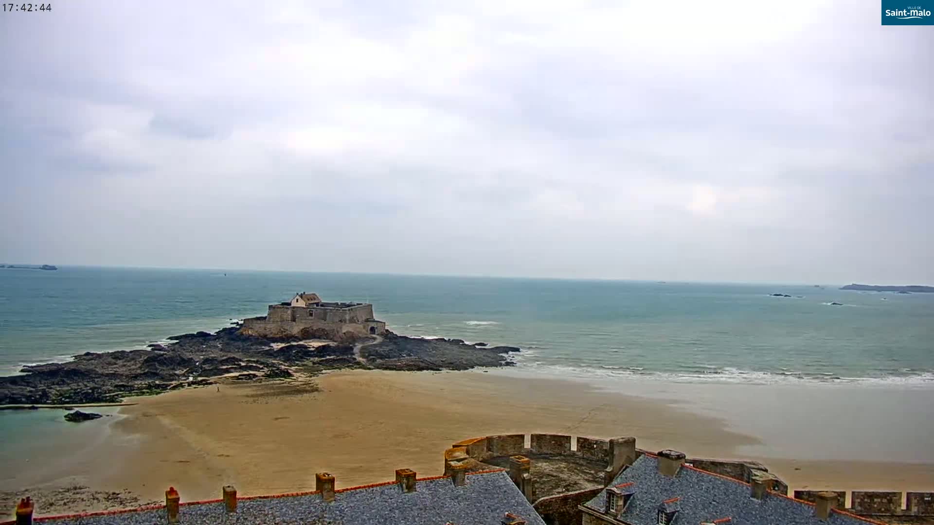A stone fortress sits on a rocky outcrop connected to a sandy beach, all under a mostly cloudy sky.