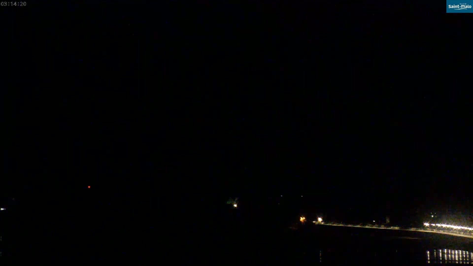 A dark nighttime scene shows a body of water reflecting a few distant lights on the opposite shore under a clear, black sky.