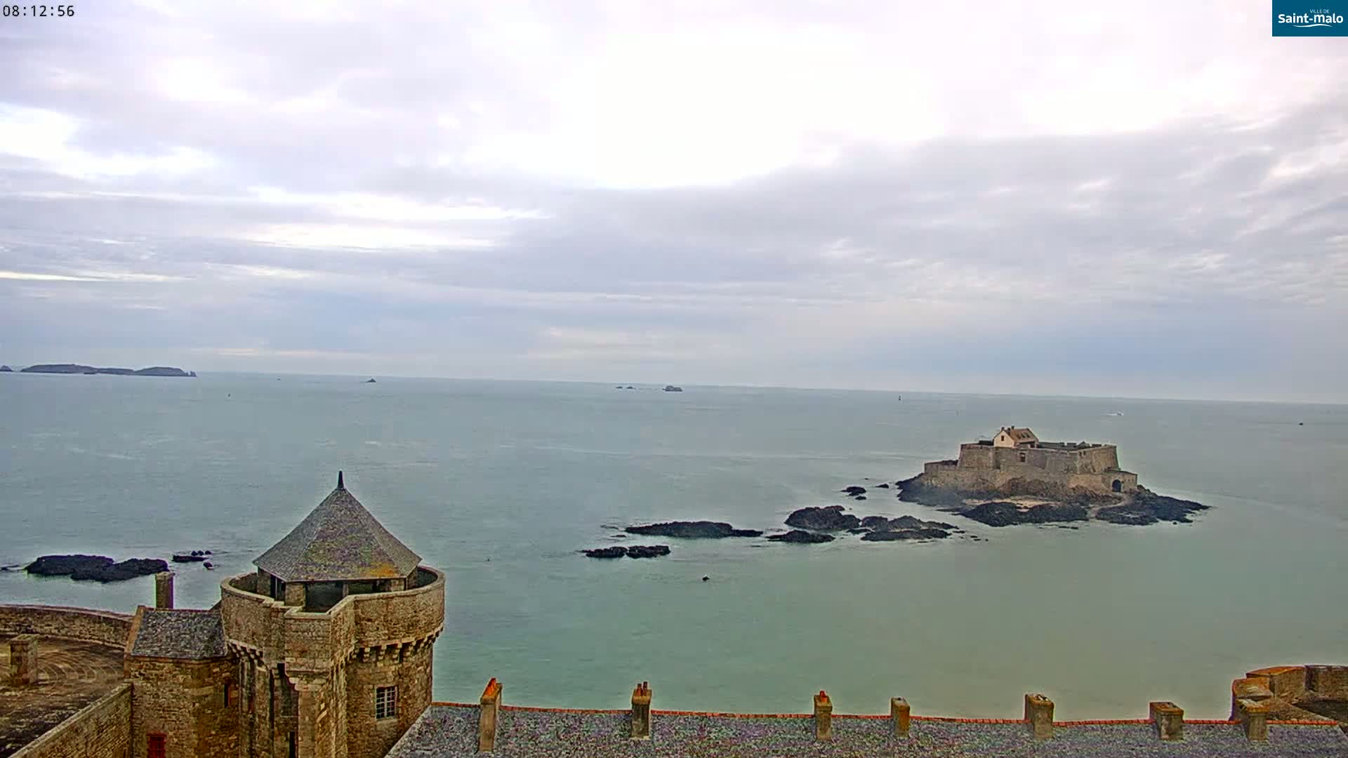 A stone castle tower overlooks a calm sea, under a cloudy sky, with a smaller fortified island visible in the distance.