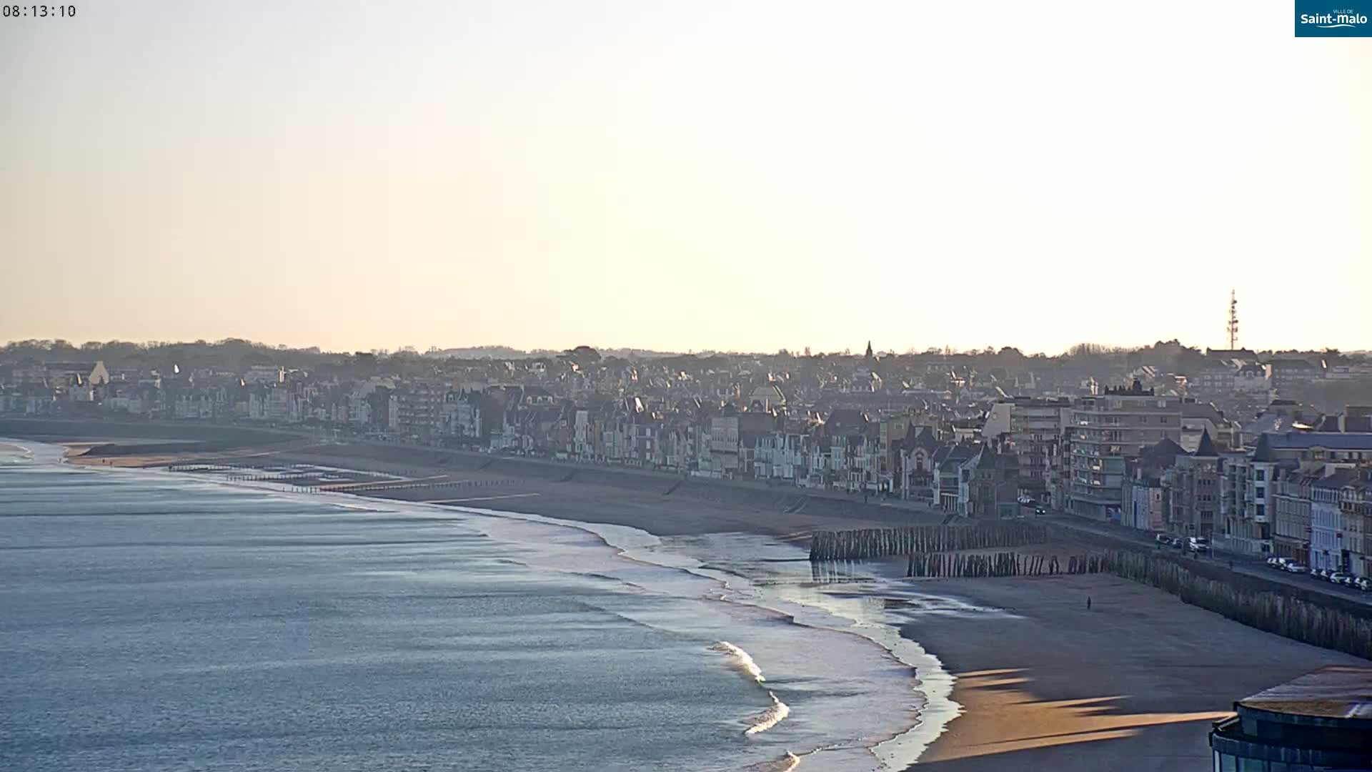 A coastal town with many buildings lines a sandy beach with gentle waves under a bright, clear sky.
