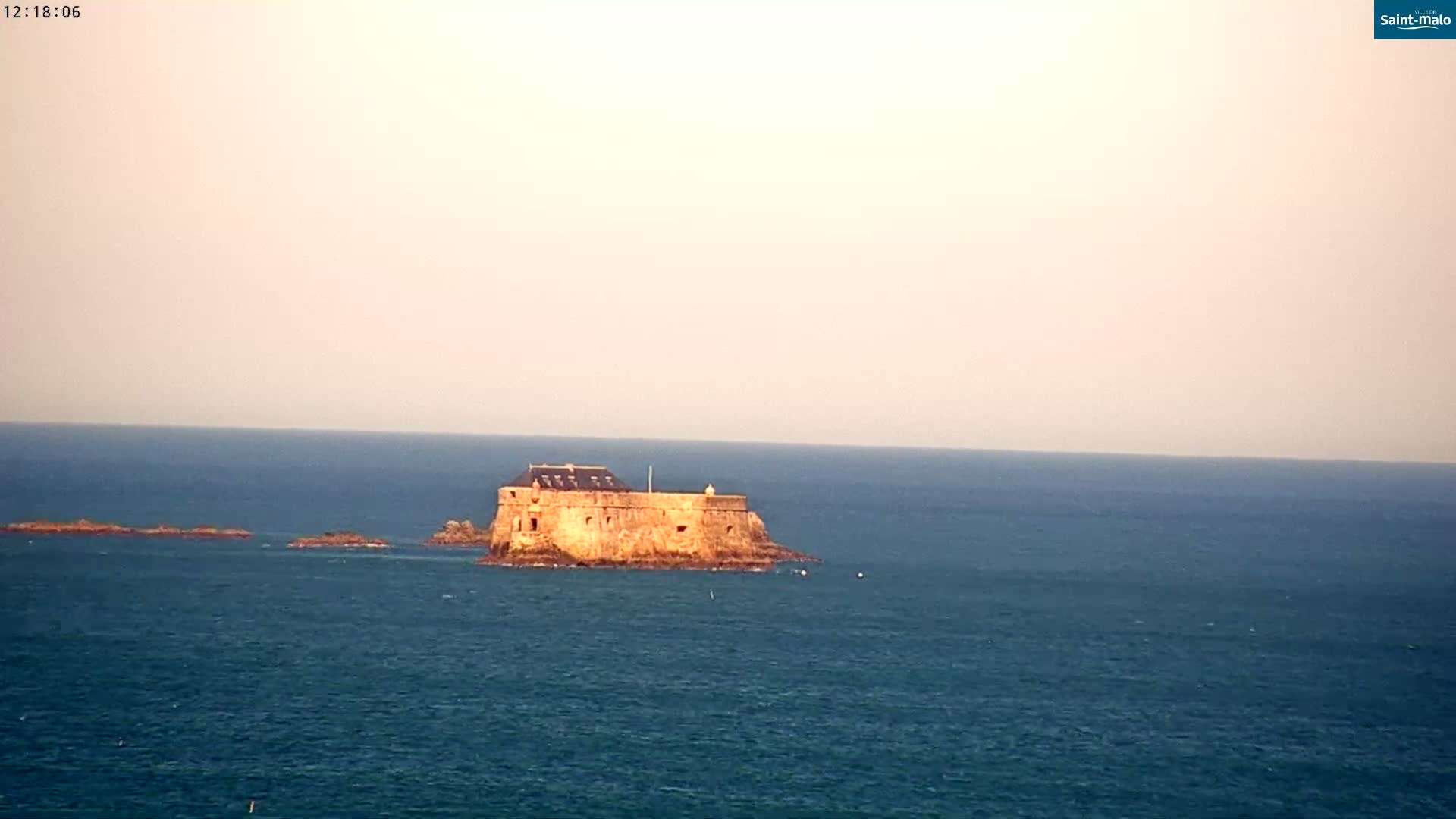 A stone fortress sits on a small island in a calm, blue ocean under a pale sky.
