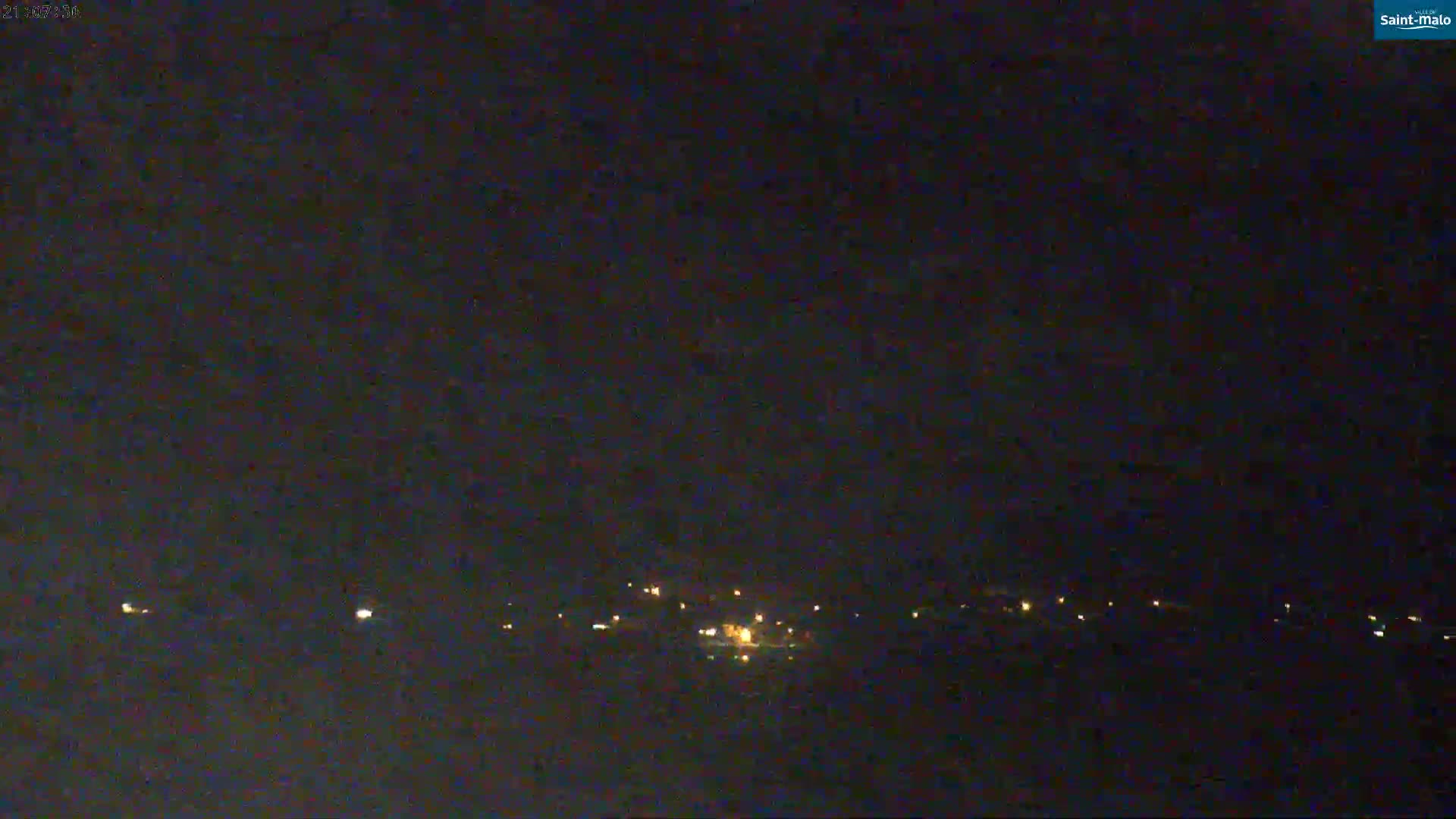 A nighttime long shot shows a cluster of faint, distant lights on a dark horizon under a clear, dark sky.