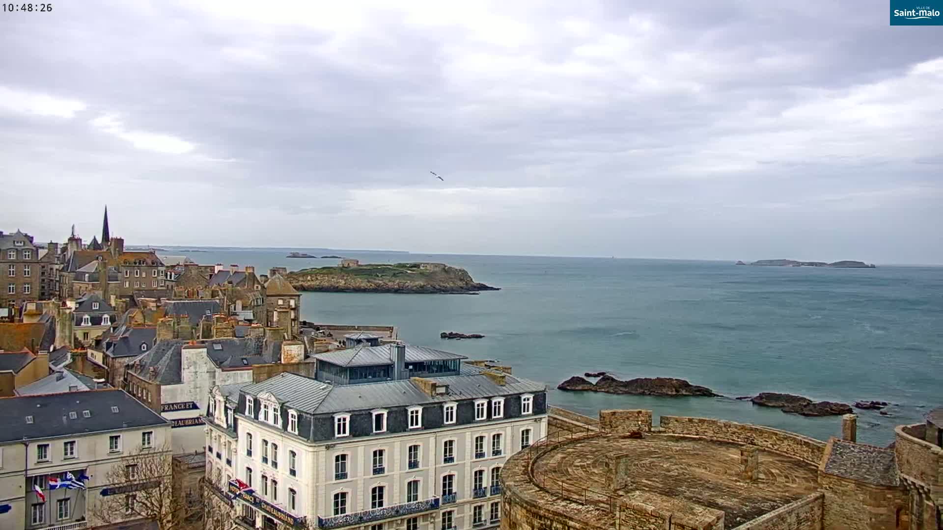 A coastal town with stone buildings and a seawall overlooks a calm, grey ocean with a small island in the distance under an overcast sky.