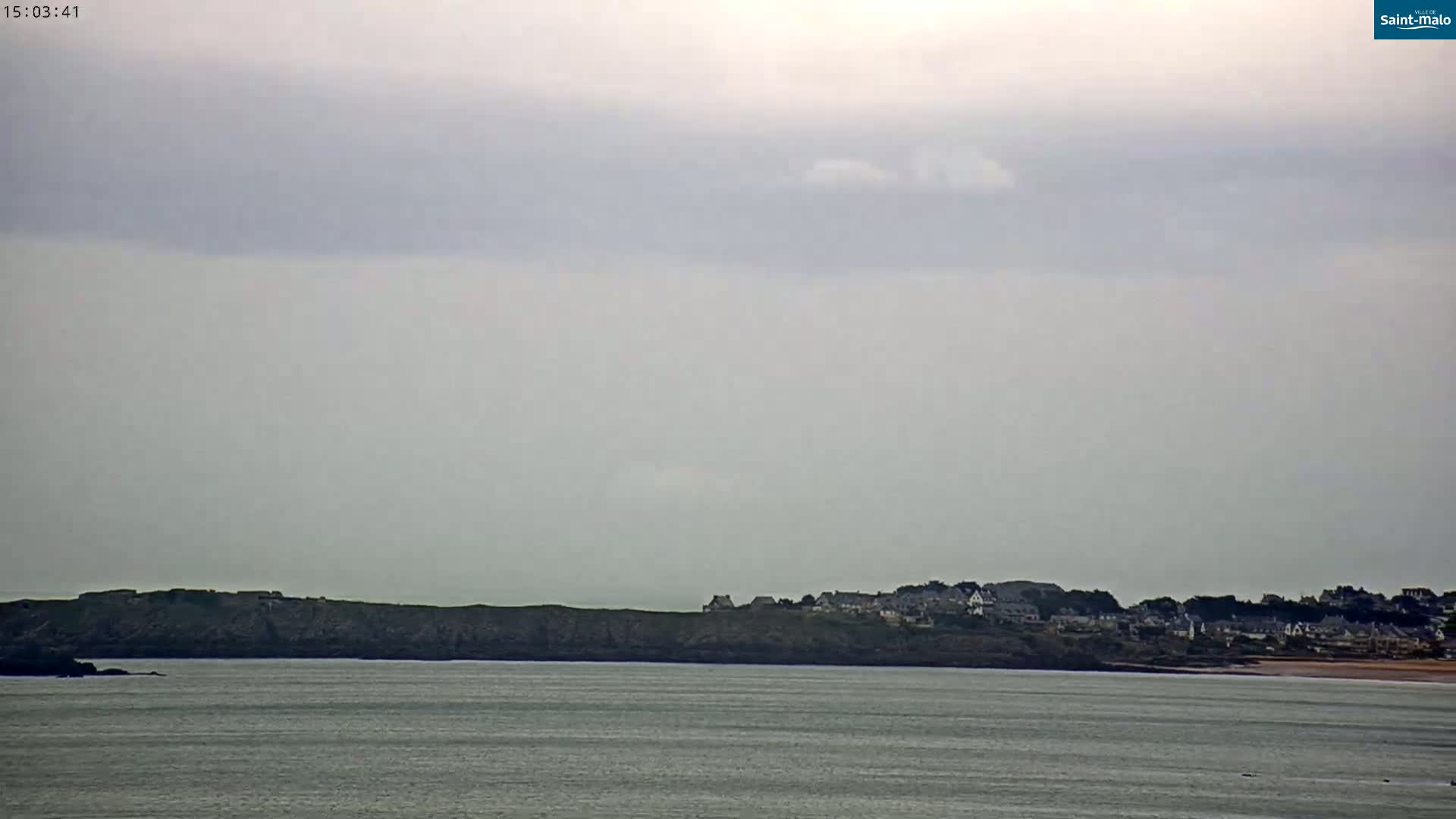 A coastal town sits atop a cliff overlooking a calm body of water under an overcast sky.
