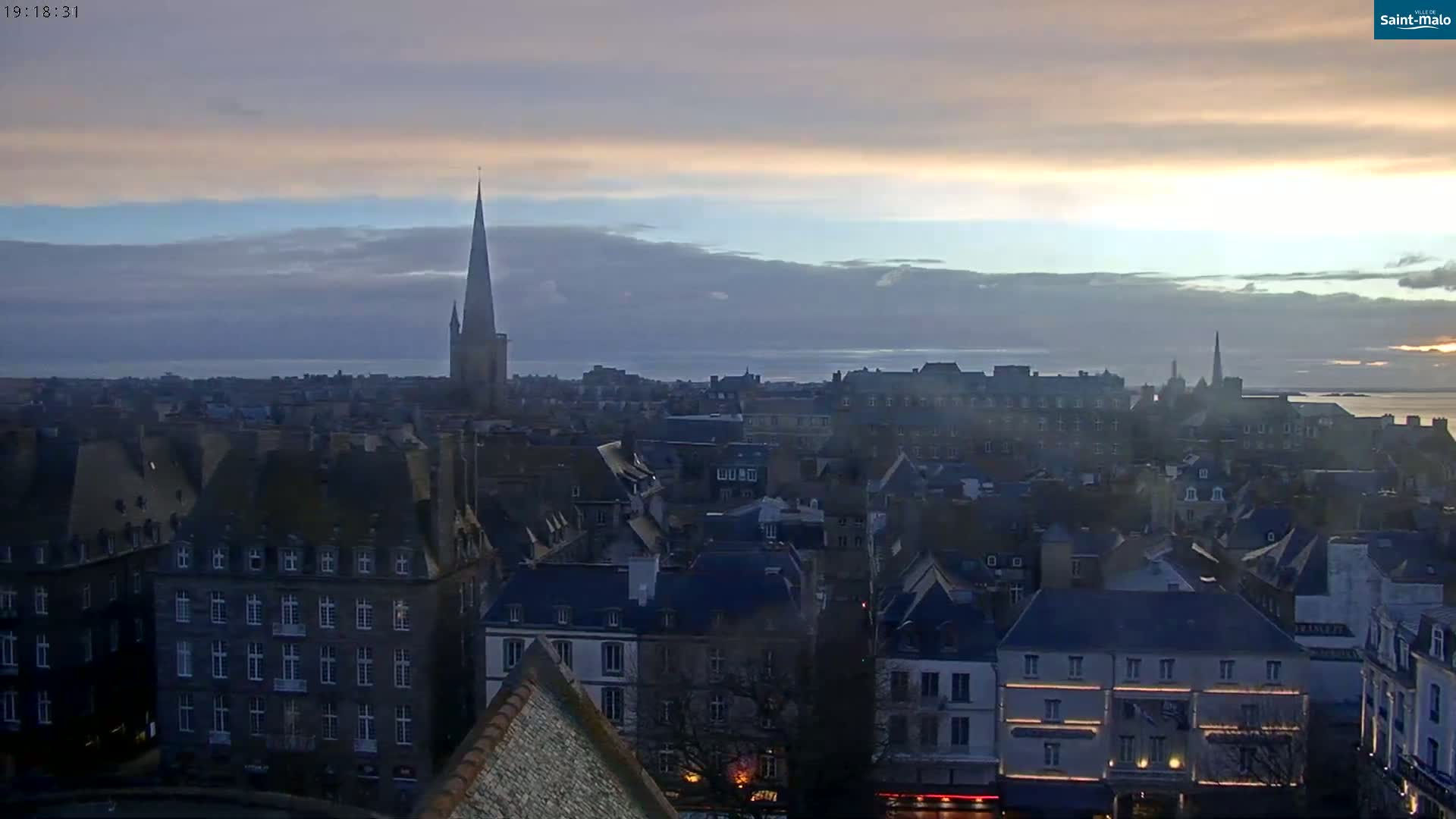 An elevated view of a European city at sunset, under a mostly cloudy sky, shows numerous buildings with dark slate roofs and a prominent church spire.