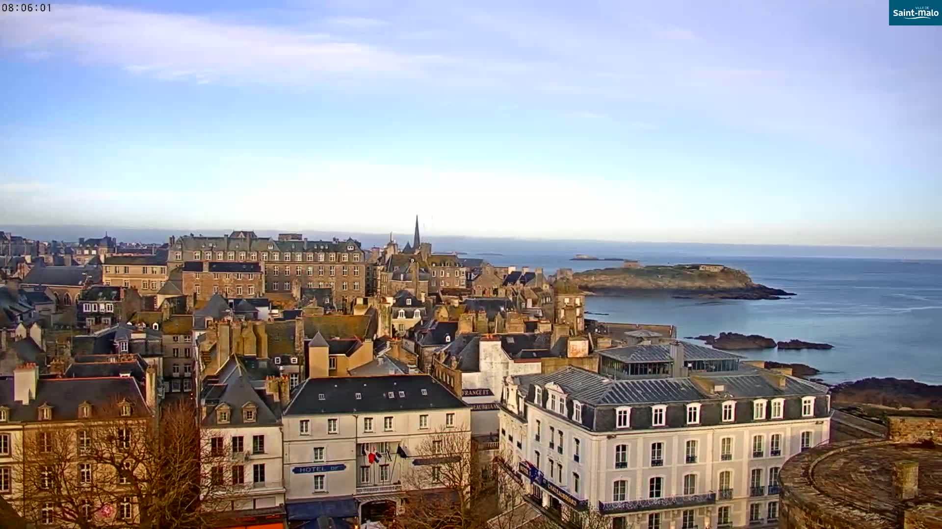 A coastal town with many stone buildings, some with visible windows and roofs, overlooks a calm ocean under a clear, bright sky.
