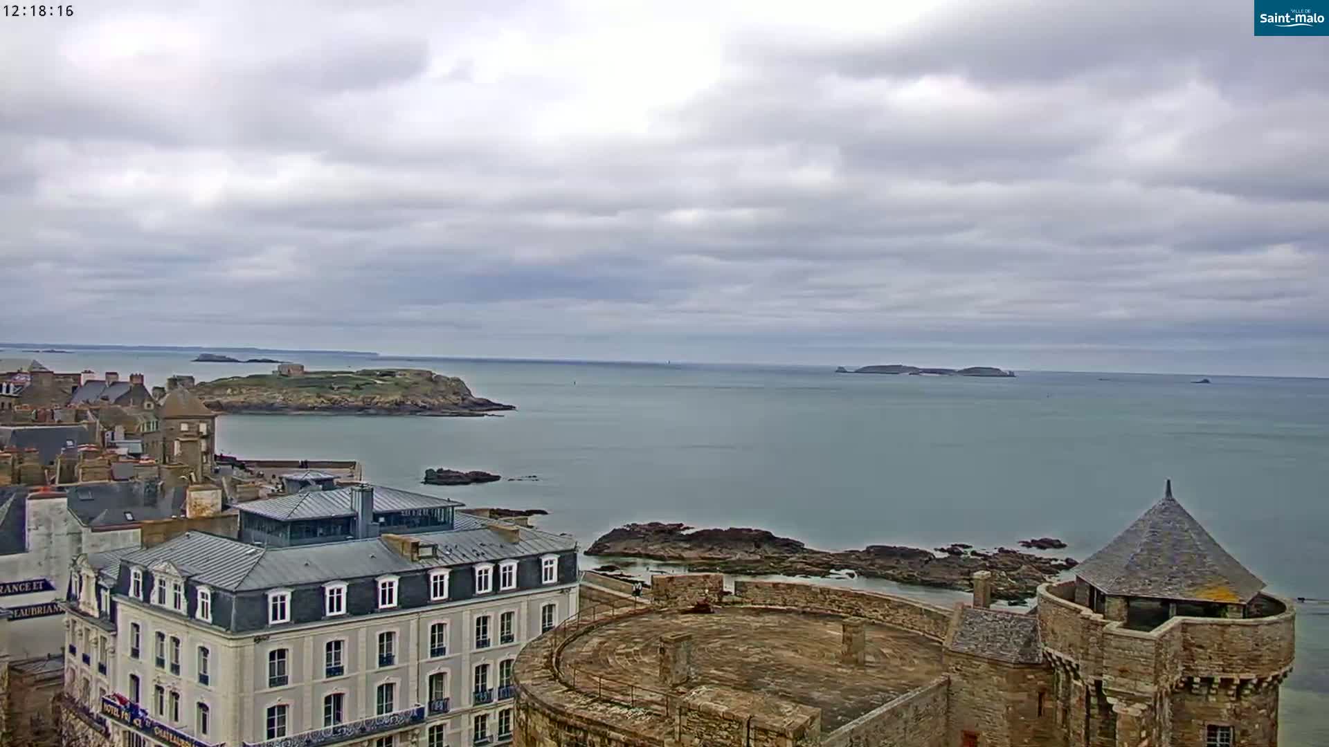 A coastal town under a cloudy sky overlooks a calm body of water with rocky shores and distant islands.