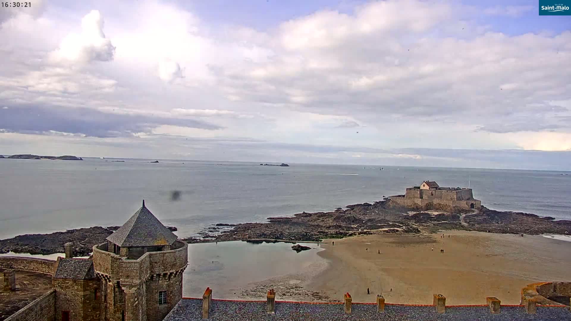 A coastal scene under a partly cloudy sky shows a stone fortress and a small beach with a few people, overlooking a calm ocean and distant islands.