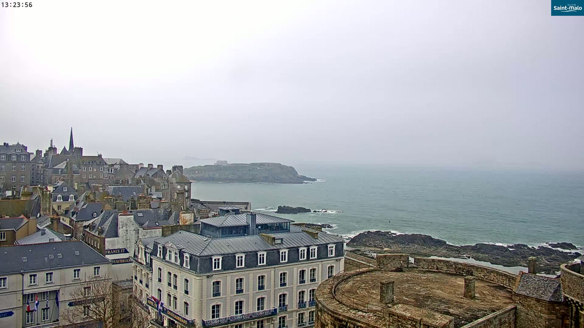 A coastal city with stone buildings overlooks a calm sea under an overcast sky.