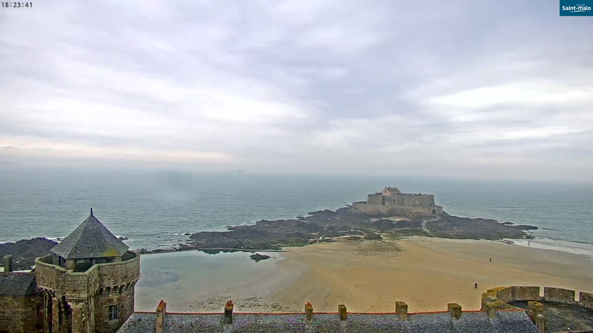 A stone castle overlooks a sandy beach and rocky coastline, with a second castle visible in the distance under a cloudy sky.