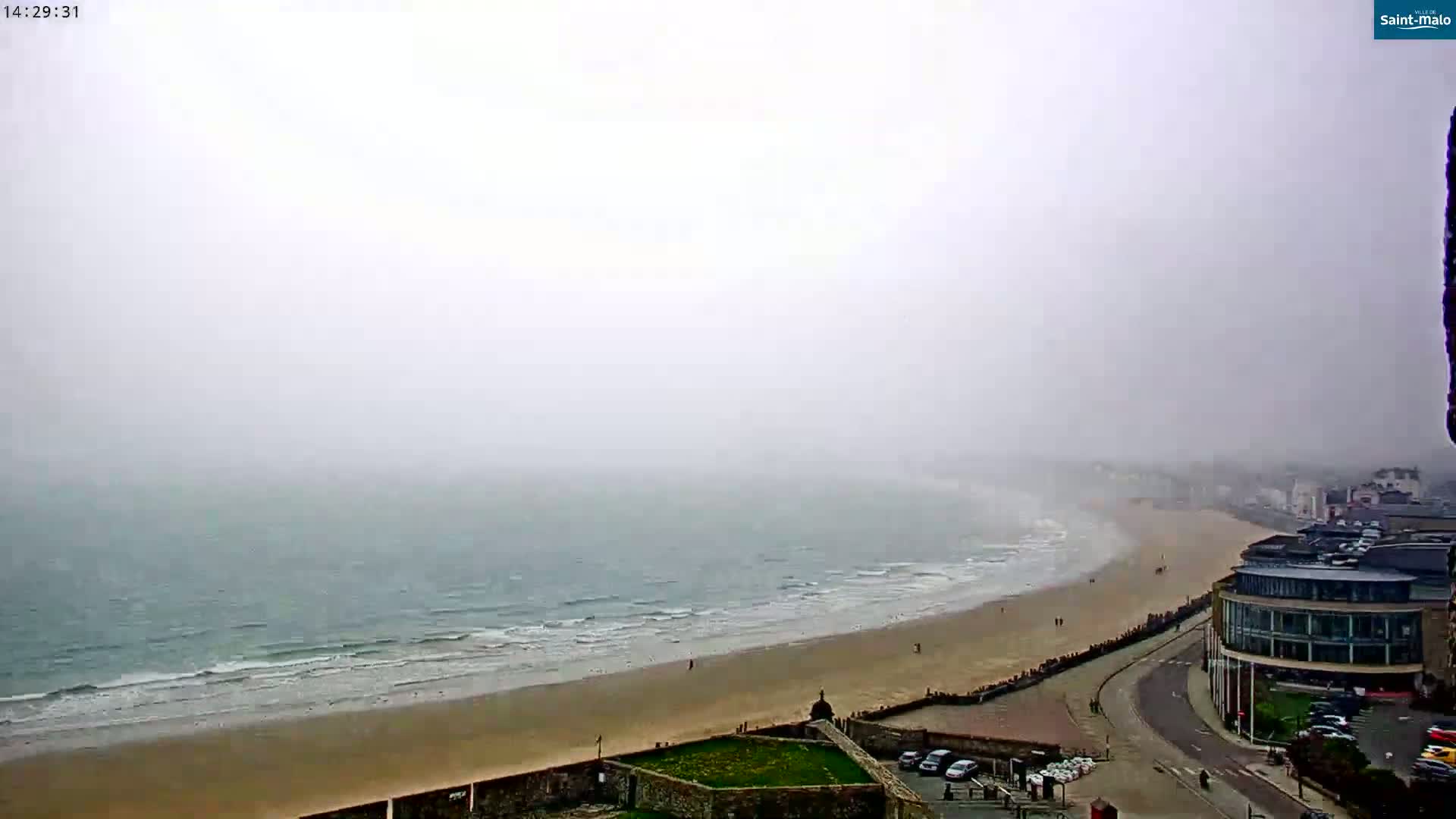 A foggy coastal scene shows a sandy beach meeting a wavy ocean under an overcast sky, with a town visible in the background.