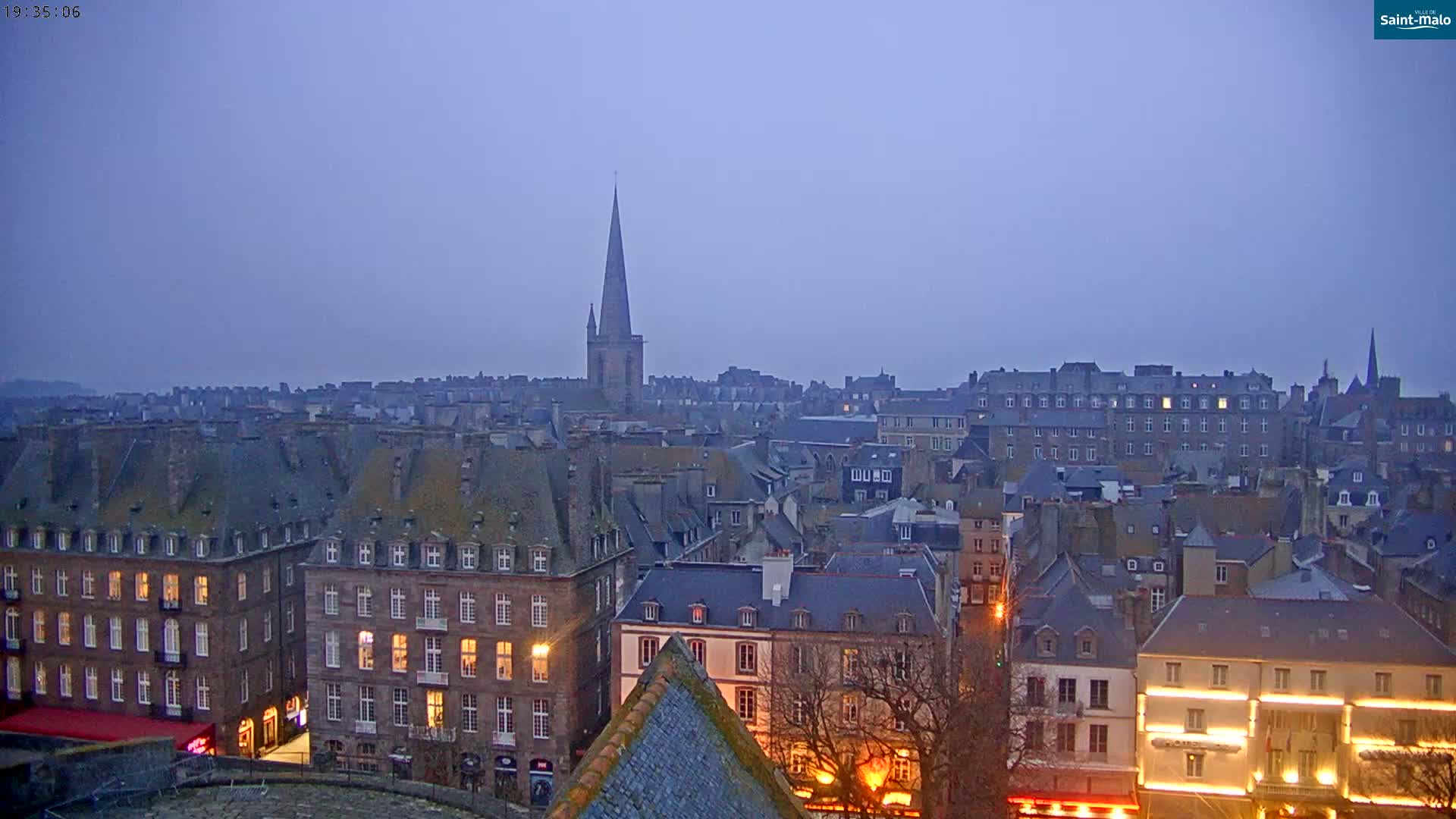 A twilight view of a European city with many dark-roofed buildings, illuminated windows, and a tall church spire in the distance under a dusky sky.