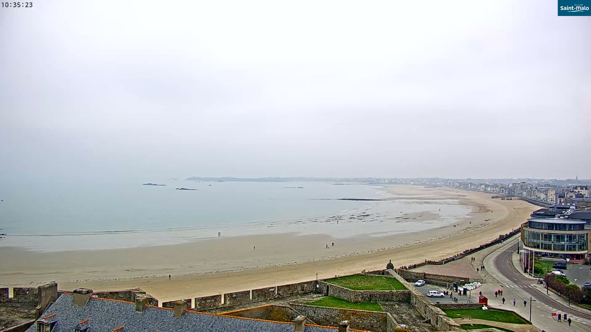 A long, sandy beach stretches along a calm, gray sea under an overcast sky, with a town visible in the distance and a stone structure in the foreground.