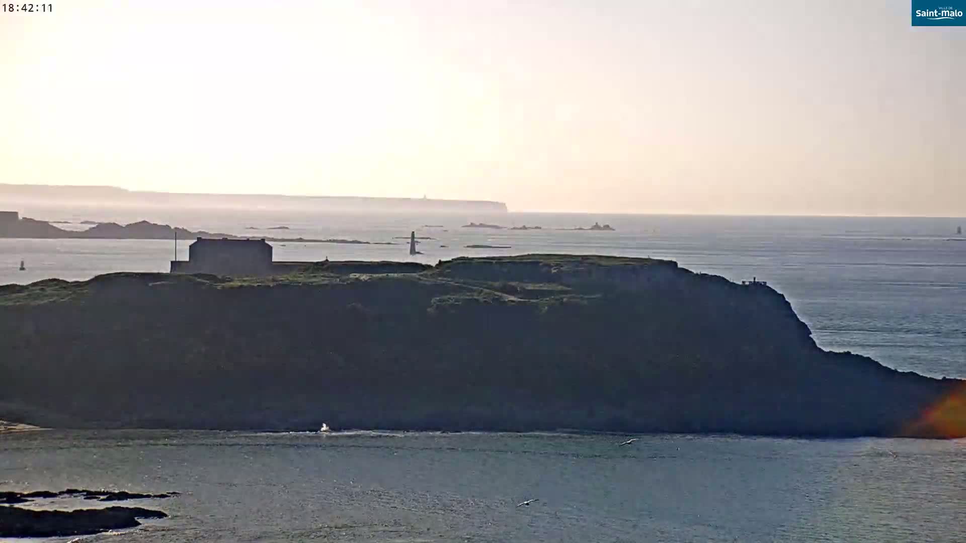 A coastal scene at sunset shows a dark, grassy cliff with a building on top, overlooking a calm sea with distant islands.