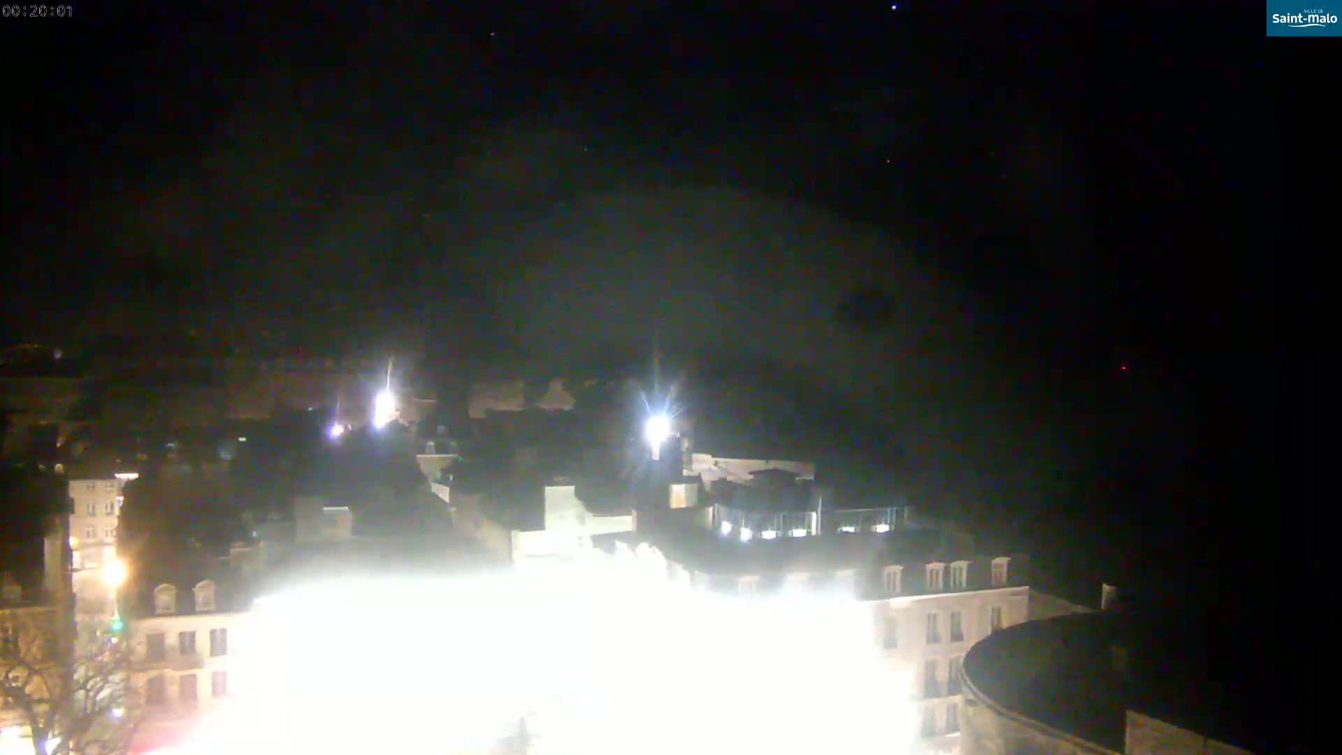 A nighttime, high-angle view shows a brightly lit town square surrounded by buildings under a dark, clear sky.