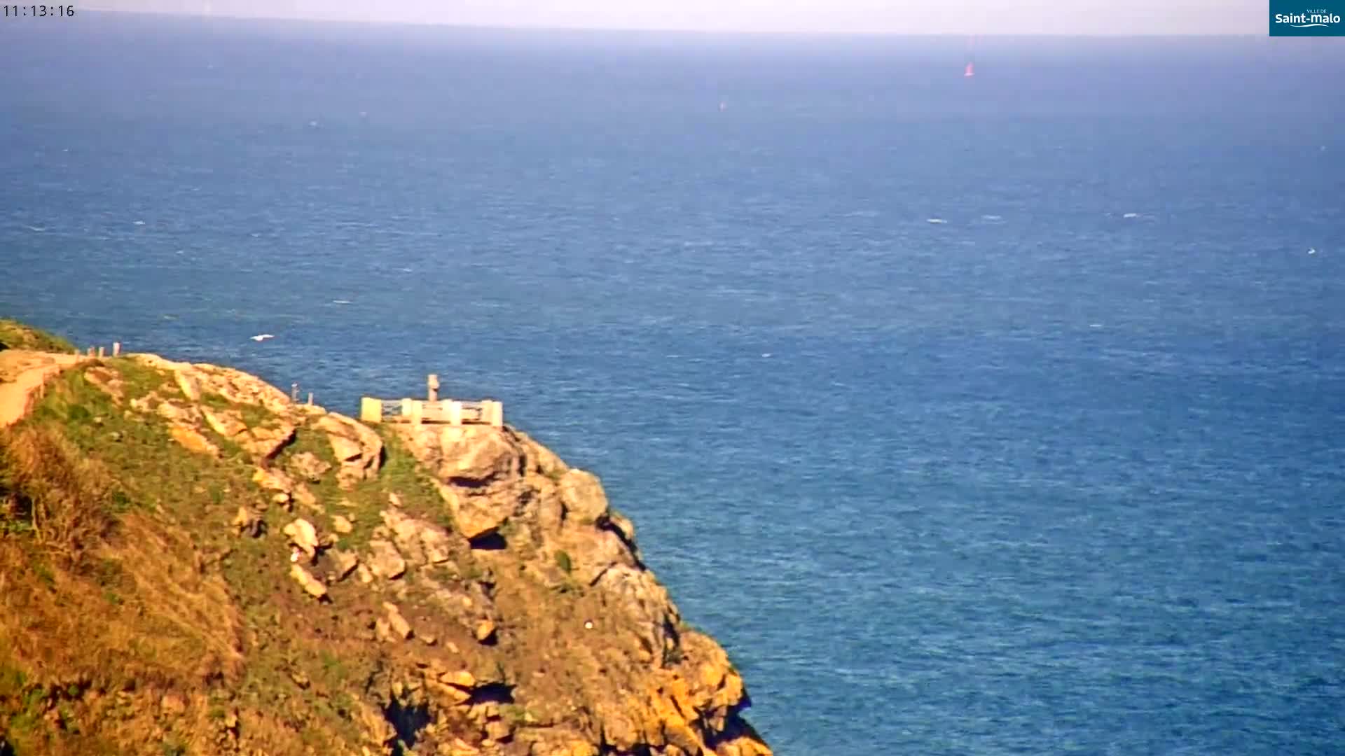 A rocky cliff overlooking a calm blue ocean on a sunny day, with a small structure atop the cliff and a path leading to it.