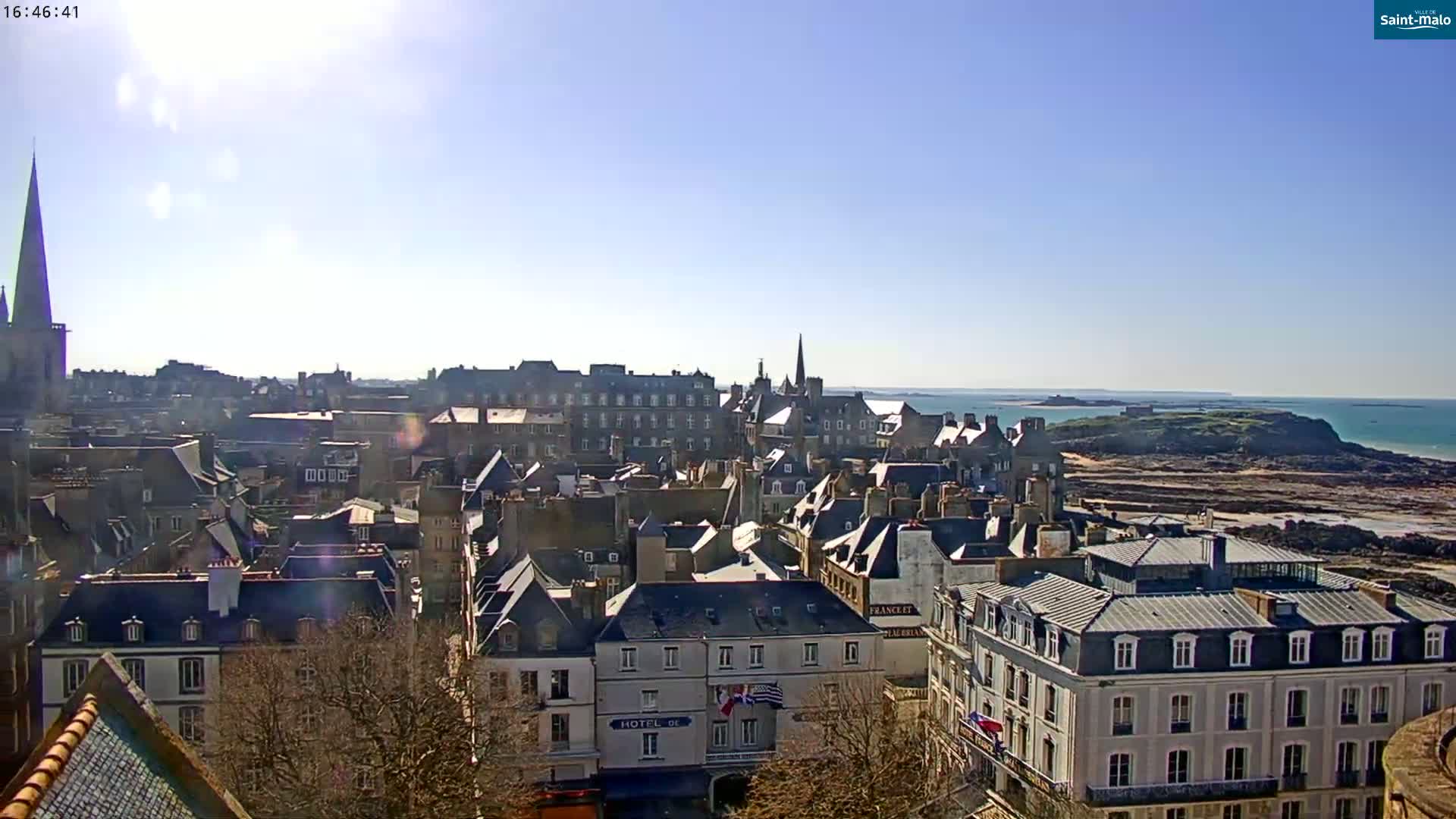 A sunny day overlooking a coastal town with a mix of architecture, including a tall church spire, and a rocky shoreline with ocean visible in the distance.