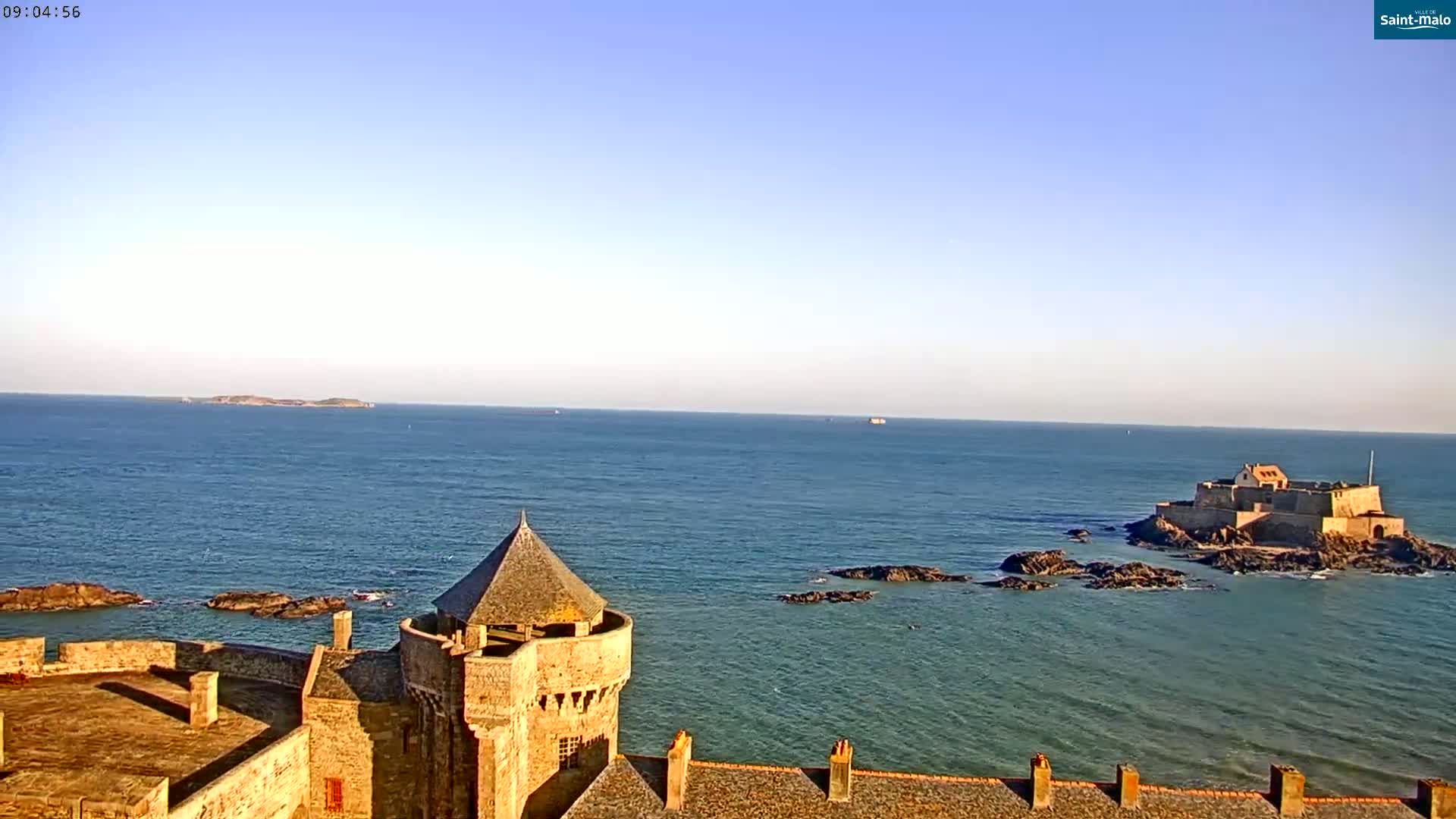 A stone tower on a coastal fortress overlooks a calm ocean, with a smaller island fortress visible in the distance under a clear sky.