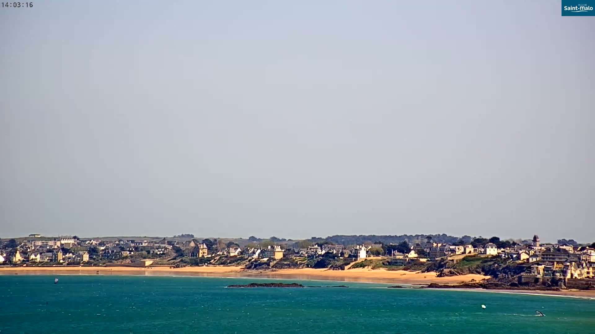 A coastal town with sandy beach stretches along a teal ocean under a hazy sky.