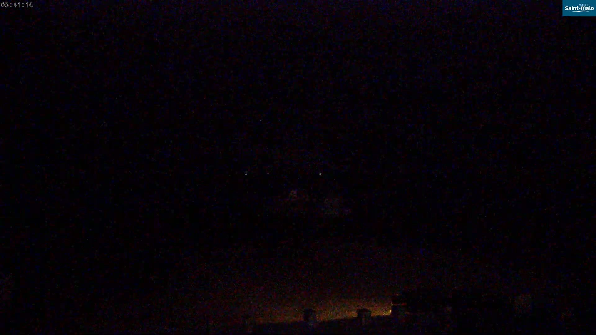A nighttime, low-light view shows a dark sky with a few faint lights and a dark horizon line with a few dim lights at the bottom.