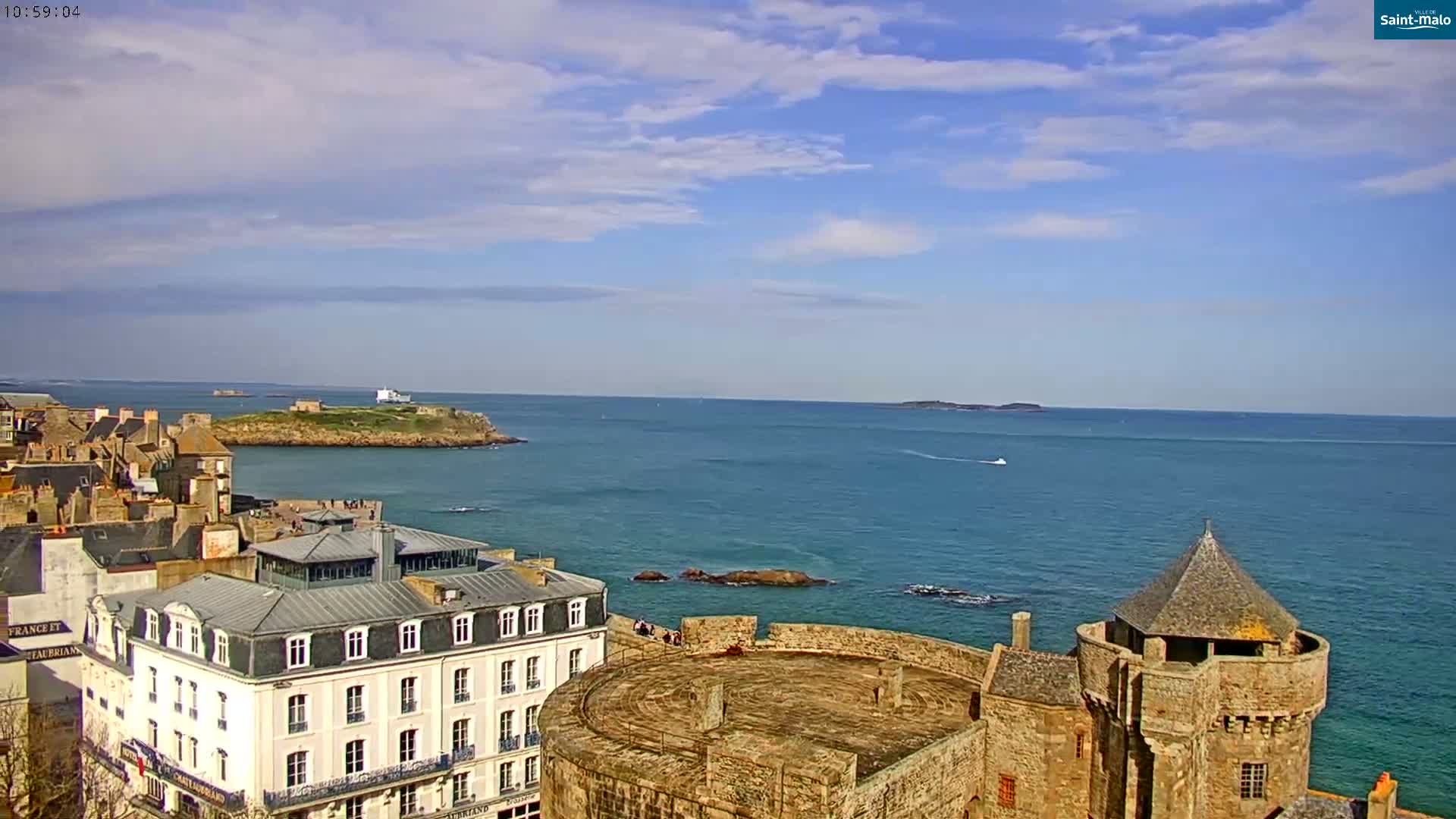 A coastal city with stone buildings and a castle overlooks a calm, blue ocean under a partly cloudy sky.